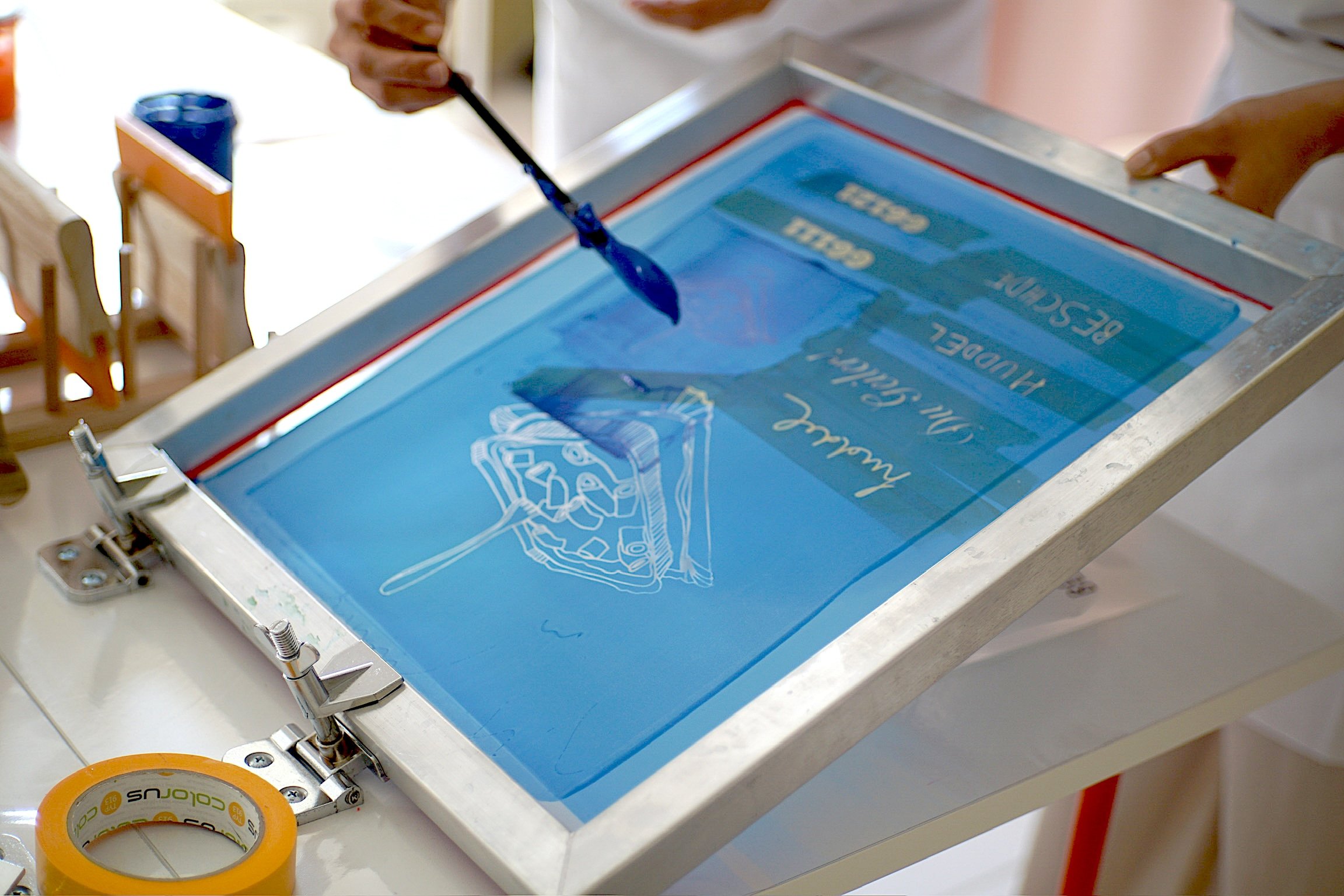 Eine Person hält einen Pinsel und arbeitet an einer Siebdruckmaschine, die ein Plakat mit blauer Hintergrundfarbe und weißen Texten bedruckt. Es befindet sich auch Abdeckband auf dem Tisch.