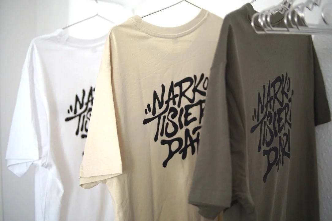 Drei T-Shirts in Weiß, Beige und Grau hängen an einer Garderobe. Jedes T-Shirt hat einen schwarzen Aufdruck mit der Aufschrift Narkotisiert in einer stilisierten, verspielten Schrift.