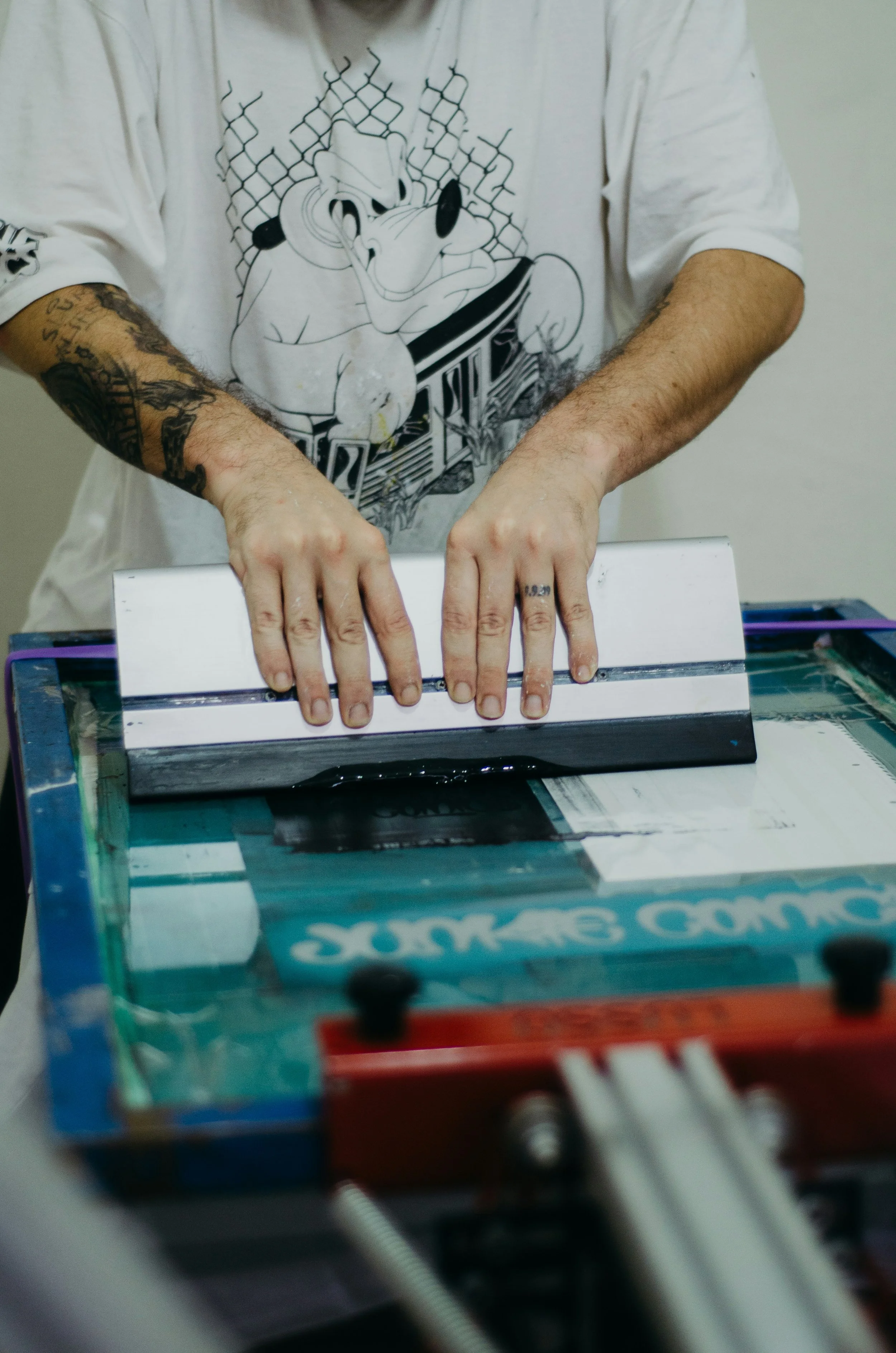 Das Siebdruck Atelier in Saarbrücken bietet Raum für kreatives Arbeiten und gemeinsames Gestalten. Person, die eine Siebdruckmaschine bedient, trägt ein weißes T-Shirt mit Textildruck.
