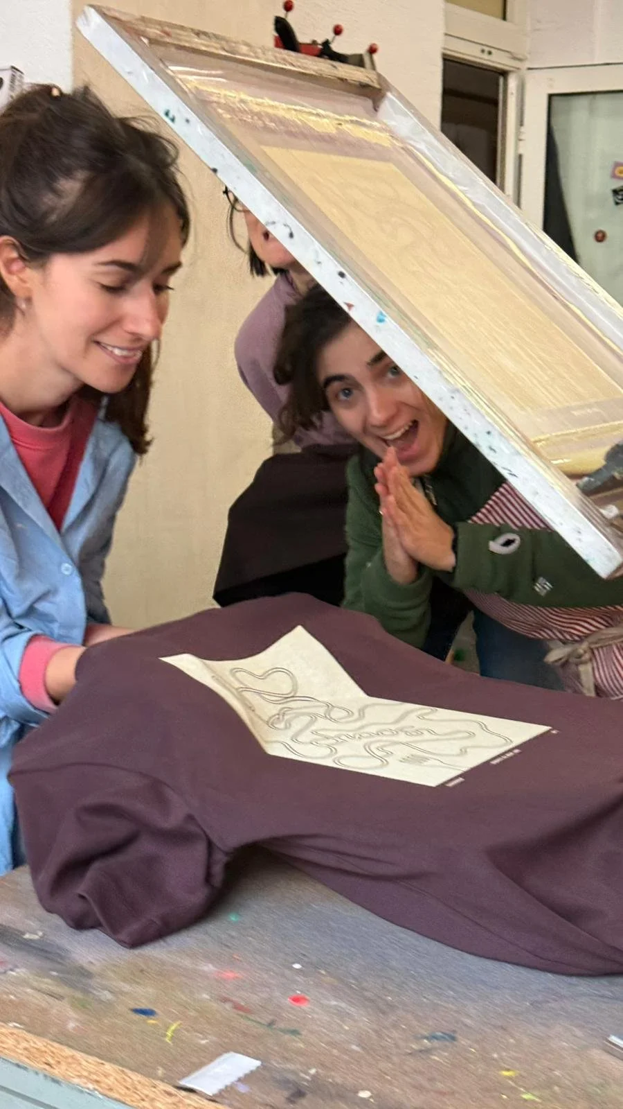 Drei Frauen unter einer Plexiglas-Box, die an einem Textildrucktisch arbeitet, lächelnd und fröhlich.