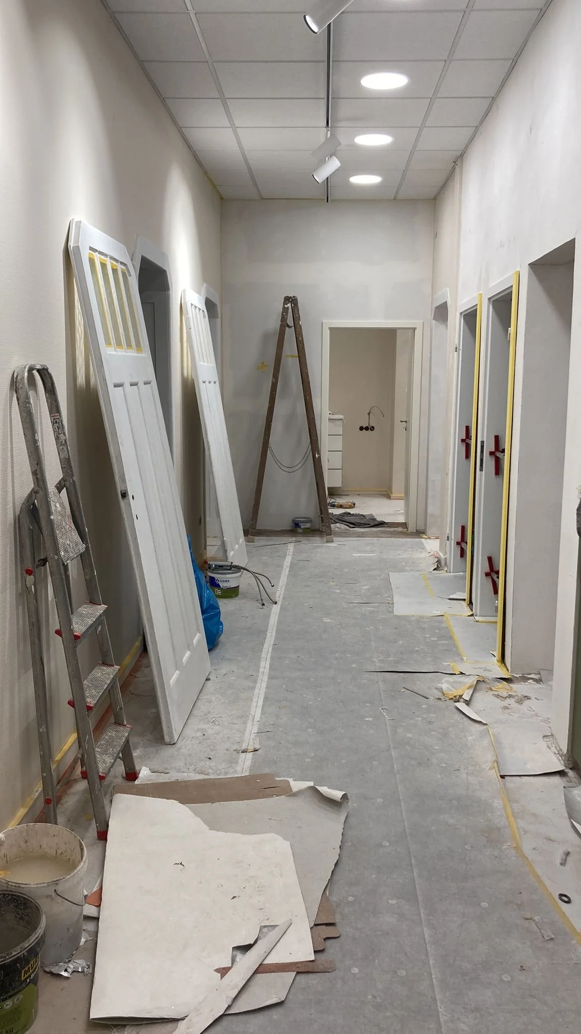 Innenaufnahme eines Flurs, der renoviert wird. Es liegen mehrere Türen gegen die Wand, einige sind bereits montiert, andere noch in Arbeit. Ein Leiter und eine kleine Trittleiter stehen im Raum. Der Boden ist mit Schutzpapier bedeckt, und verschiedene Werkzeuge und Farbdosen liegen herum.