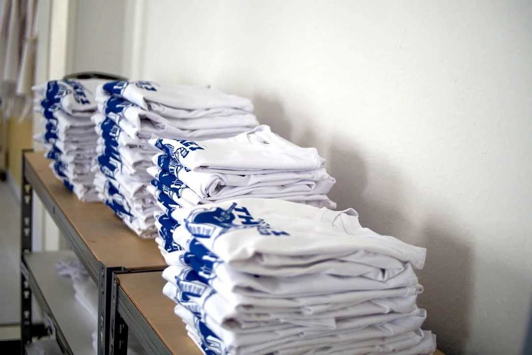 Mehrere Stapel weißer T-Shirts mit blauer Grafik liegen auf einem Tisch.