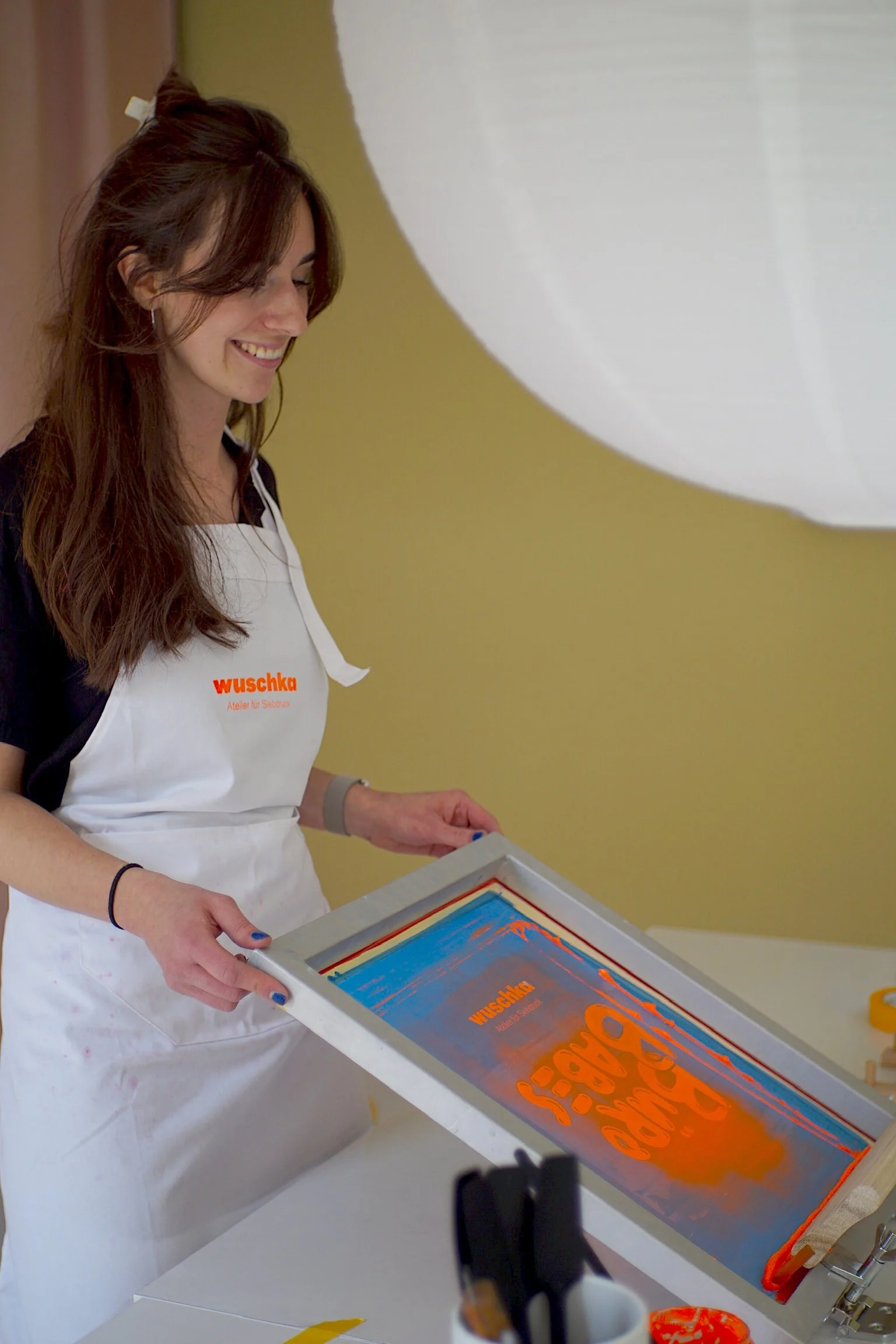 Silvia Wilhelm trägt eine weiße Schürze mit dem Logo 'wuschka' und hält eine Druckplatte in der Hand, die künstlerische, bunte Drucke zeigt. Sie lächelt und steht vor einem gelben Hintergrund.