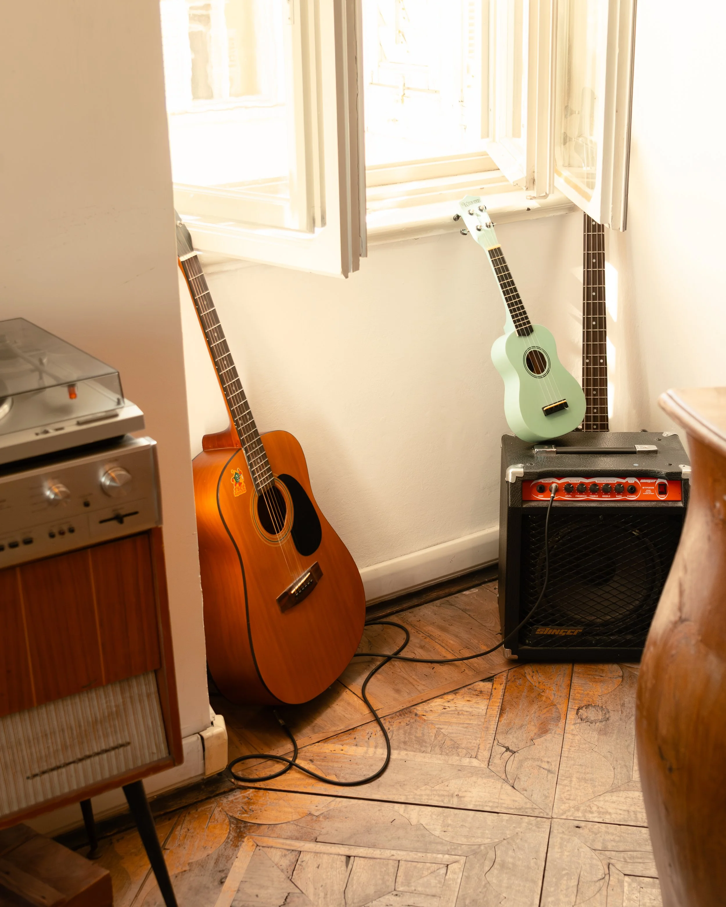 Two acoustic guitars at Controvento Hostel, one wooden and one mint green, leaning against a wall with a small orange amplifier beneath the green guitar, photographed by Skye J Blount.