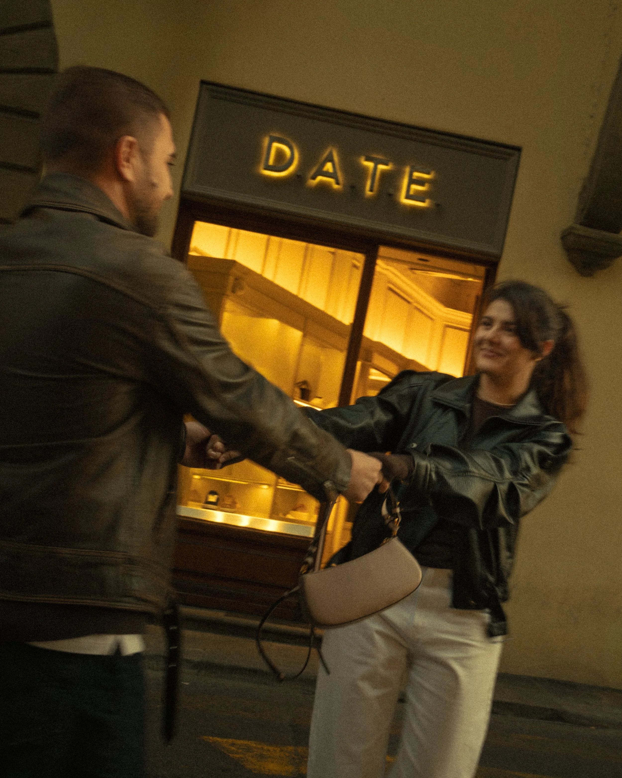 Cinematic portrait of a man and woman shaking hands outside a building with an illuminated ‘DATE’ sign, both wearing leather jackets, photographed by Skye J Blount.