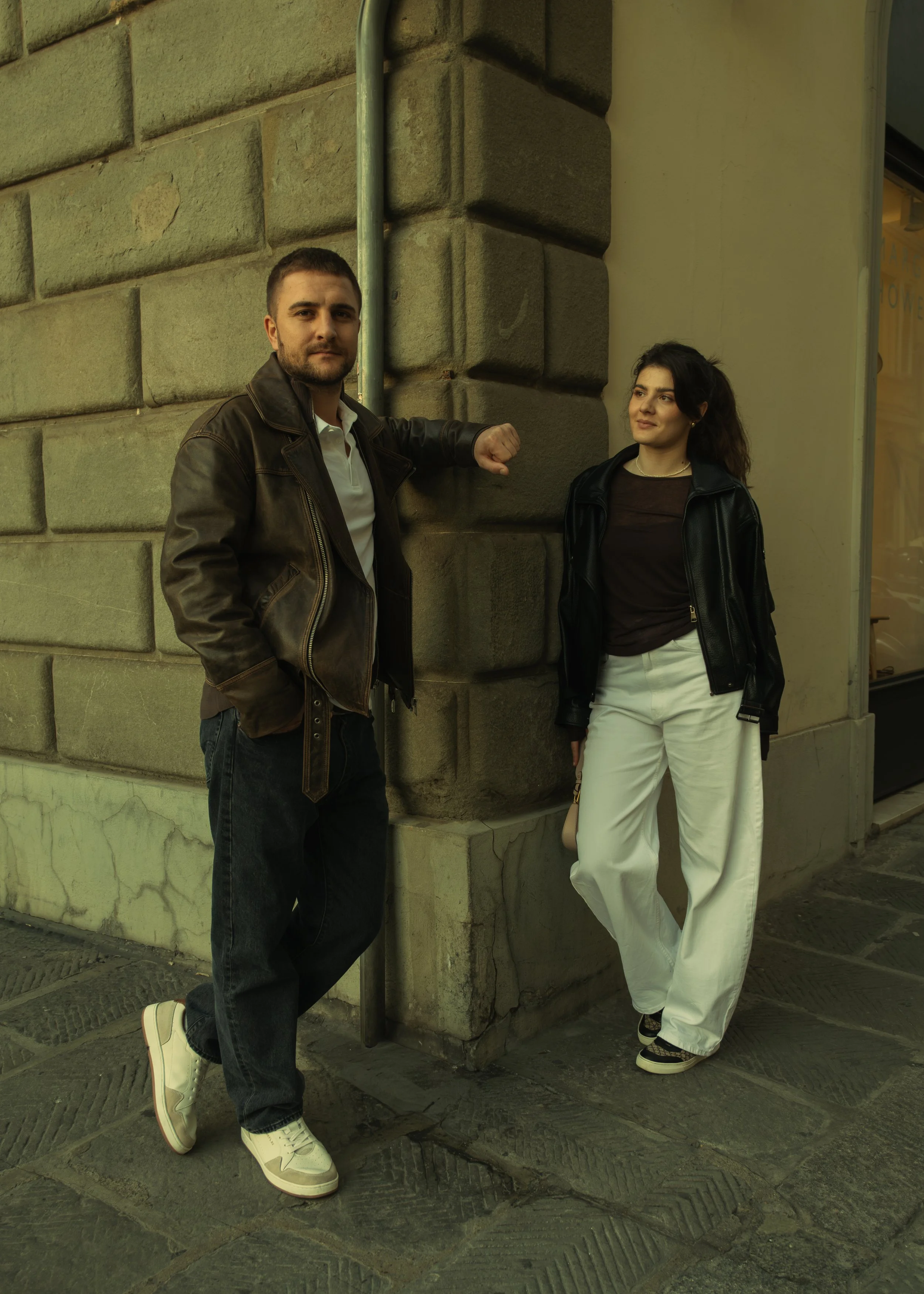 Cinematic portrait of a man and woman standing on a city street leaning against a stone wall, with the man showing a fist bump gesture and the woman standing casually, photographed by Skye J Blount.