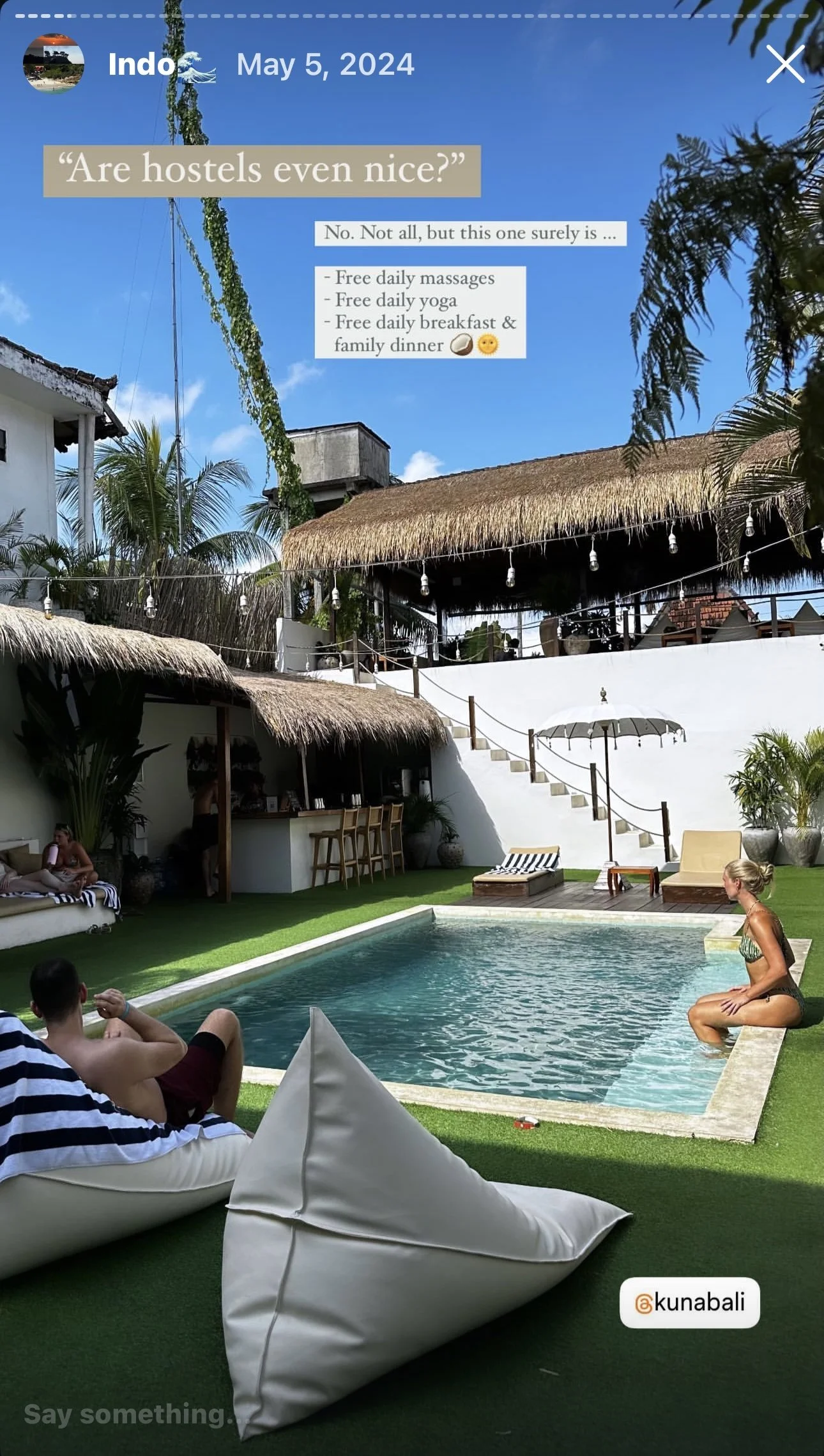 A tropical outdoor pool area with a woman sitting on the pool's edge, a man lounging on a bean bag chair, and a bar under a thatched roof. Palm trees, a clear blue sky, and string lights are visible, creating a relaxing vacation vibe.