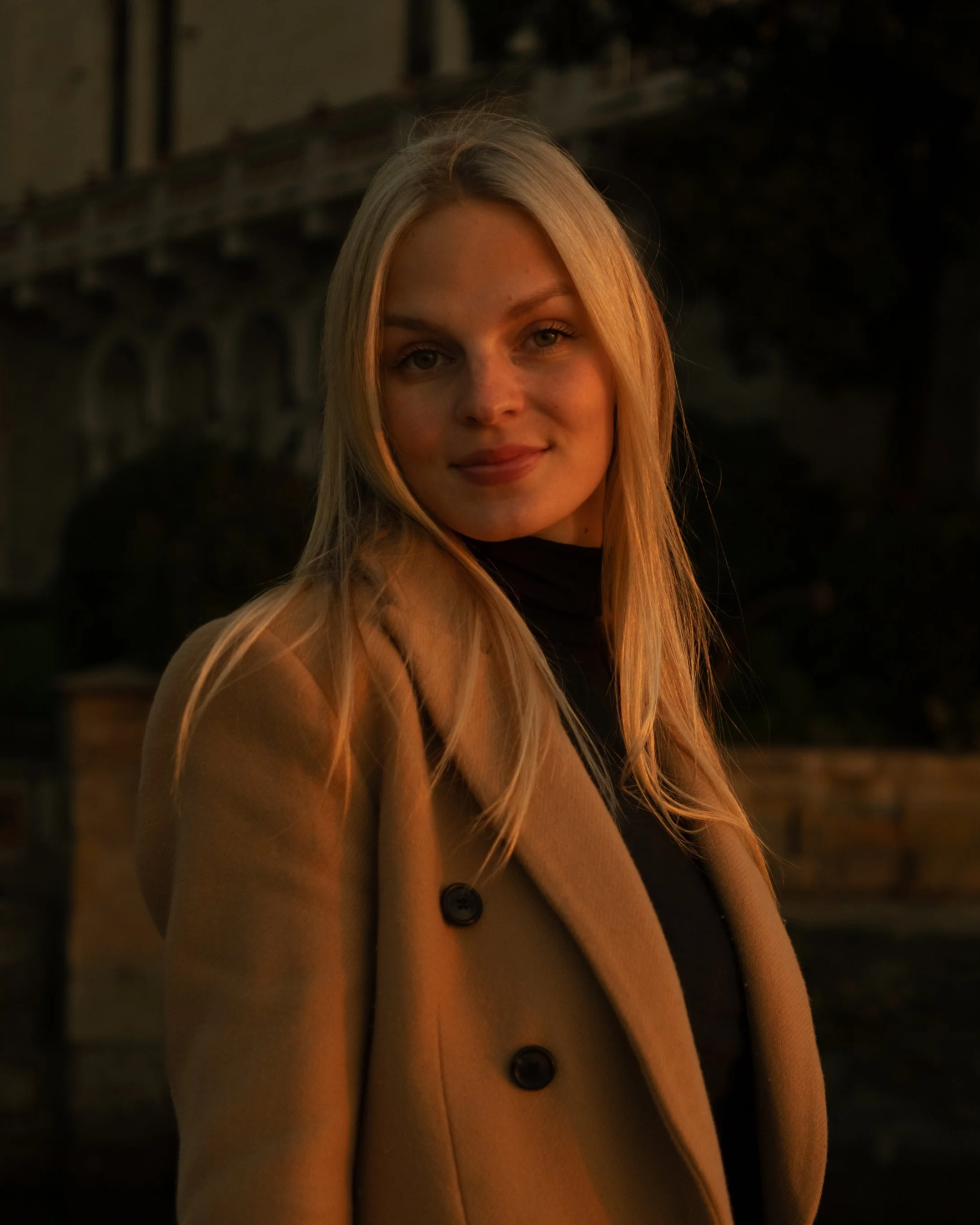 Portrait of a young blonde woman wearing a tan coat and black turtleneck, smiling outdoors at dusk, photographed by Skye J Blount.