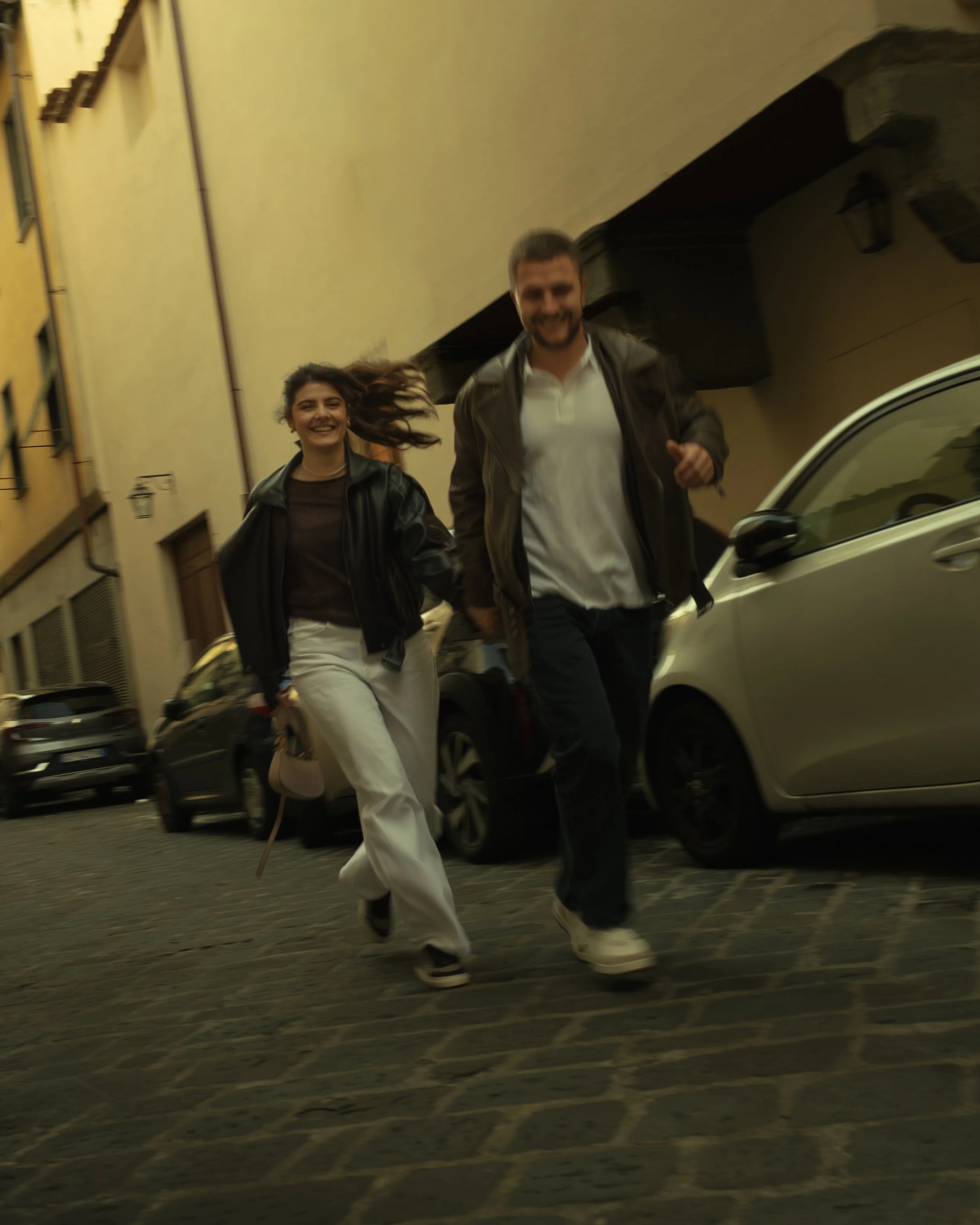Cinematic portrait of a happy couple running together on a cobblestone street in Florence with parked cars and yellow buildings, photographed by Skye J Blount.