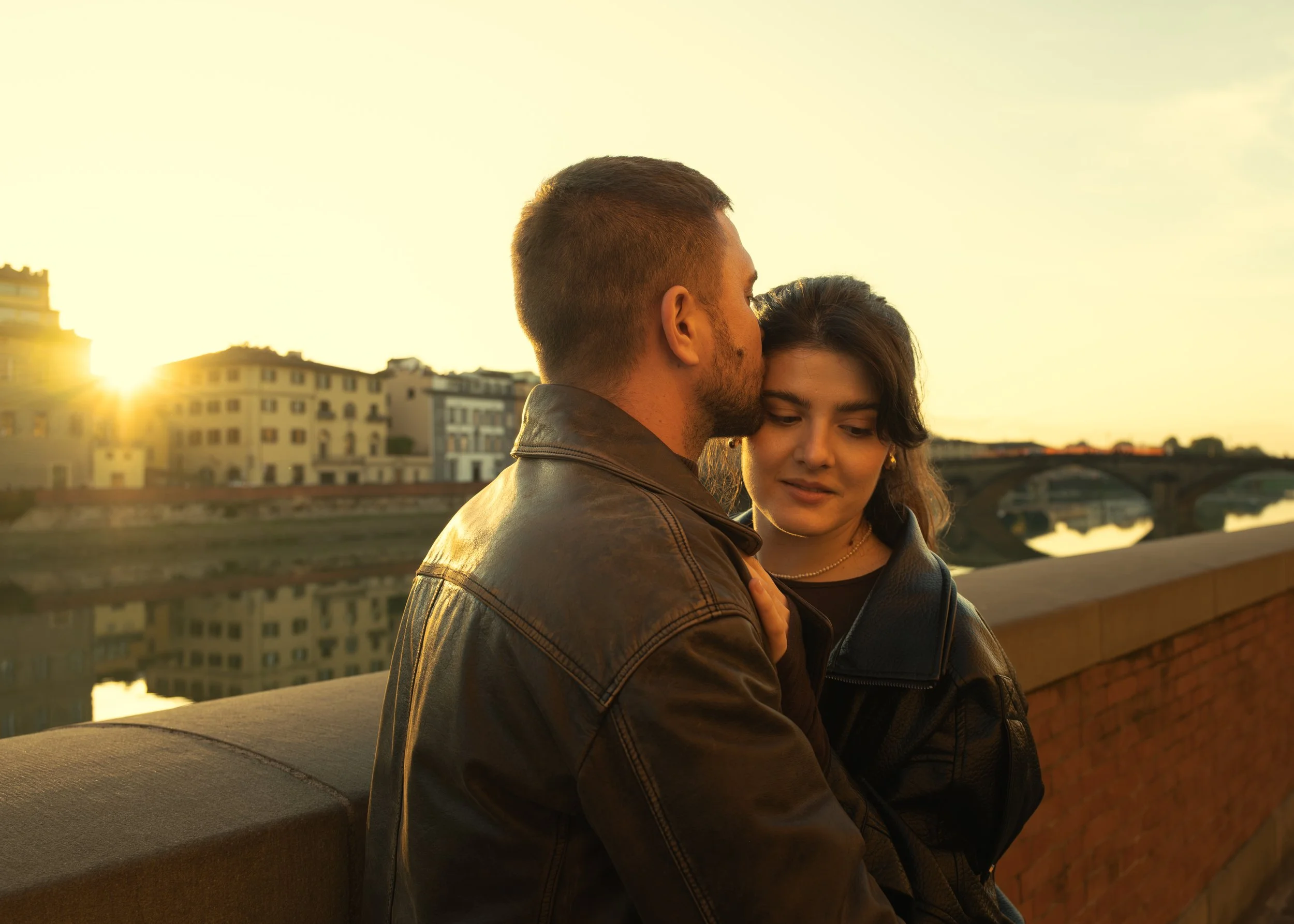 Cinematic portrait of a couple sharing an intimate moment at sunset on a bridge with a cityscape in the background, photographed by Skye J Blount.