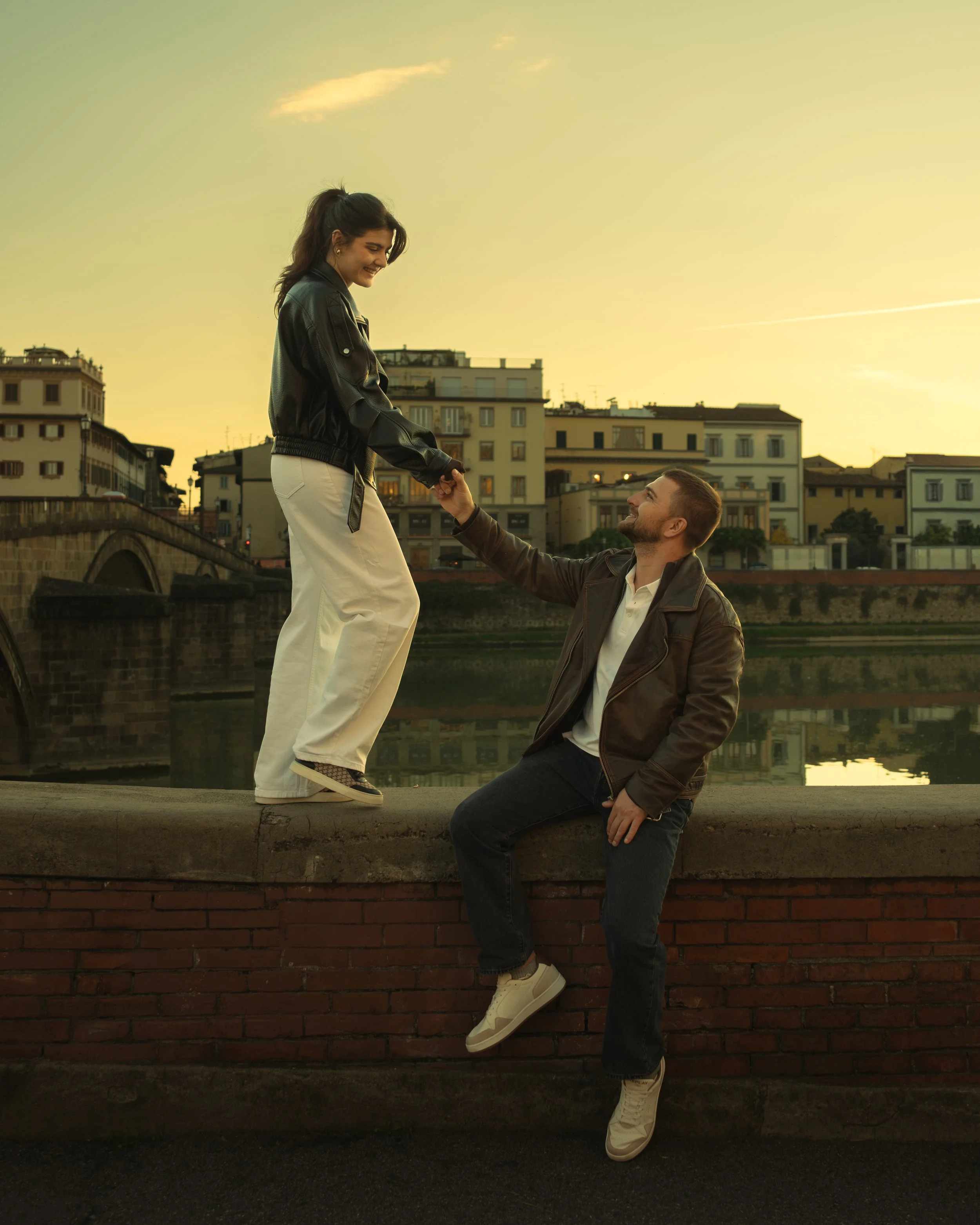 Cinematic portrait of a man helping a woman stand on the edge of a wall during sunset with city buildings in Florence in the background, photographed by Skye J Blount.