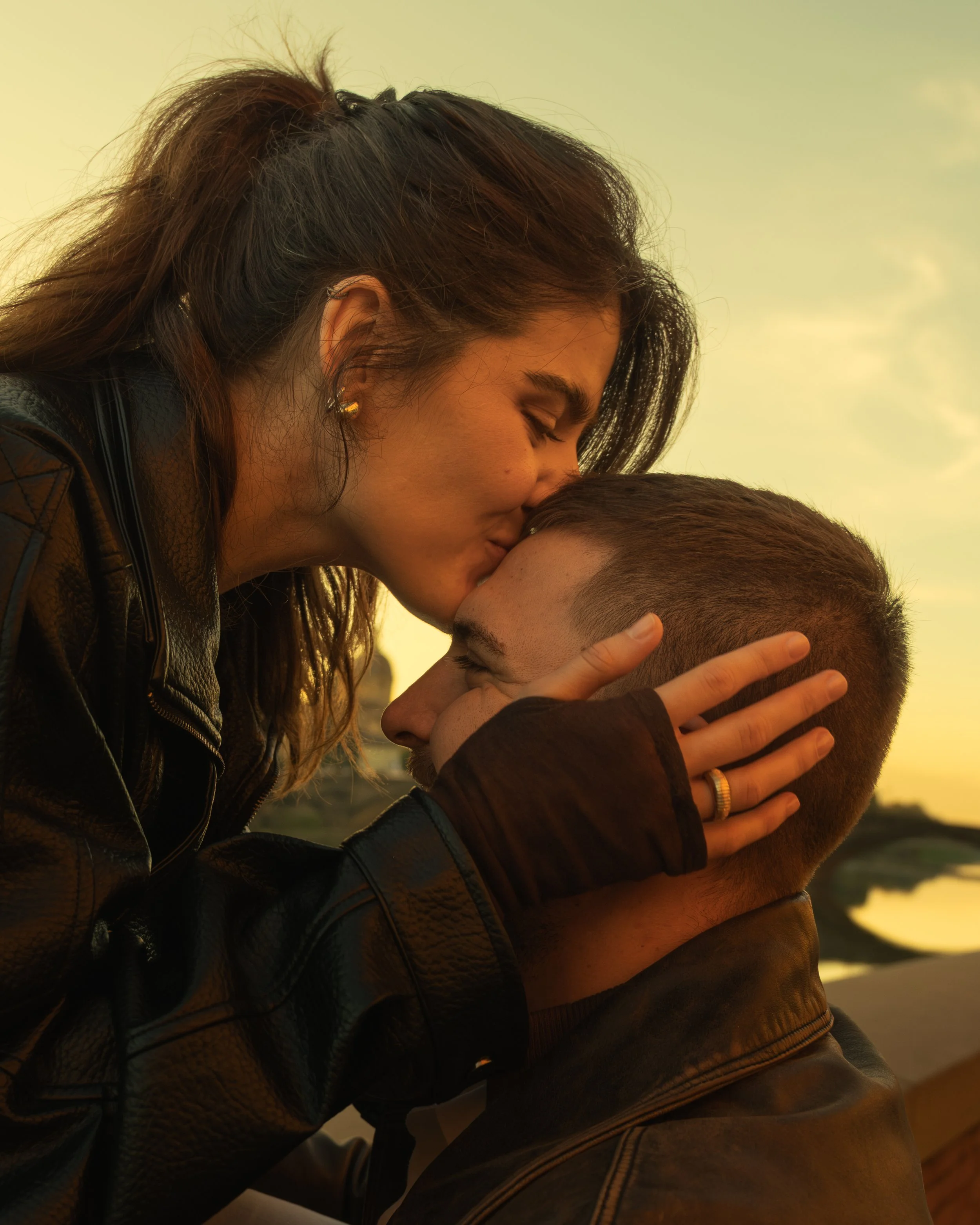 Cinematic portrait of a woman kissing a man on the forehead as he holds her face outdoors during sunset, photographed by Skye J Blount.