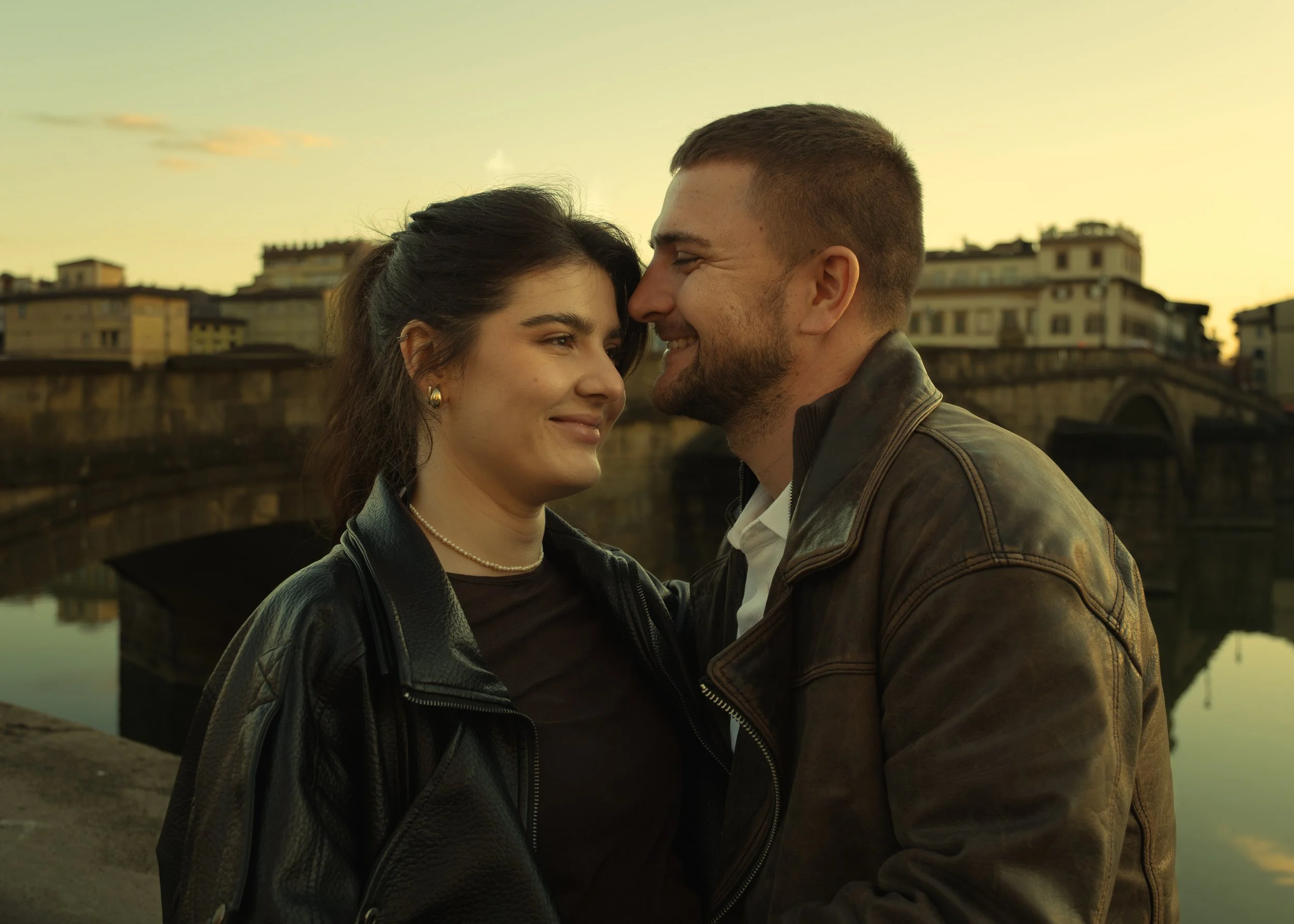 A young couple smiling and leaning close to each other near a river during sunset in an urban setting, photographed by Skye J Blount.