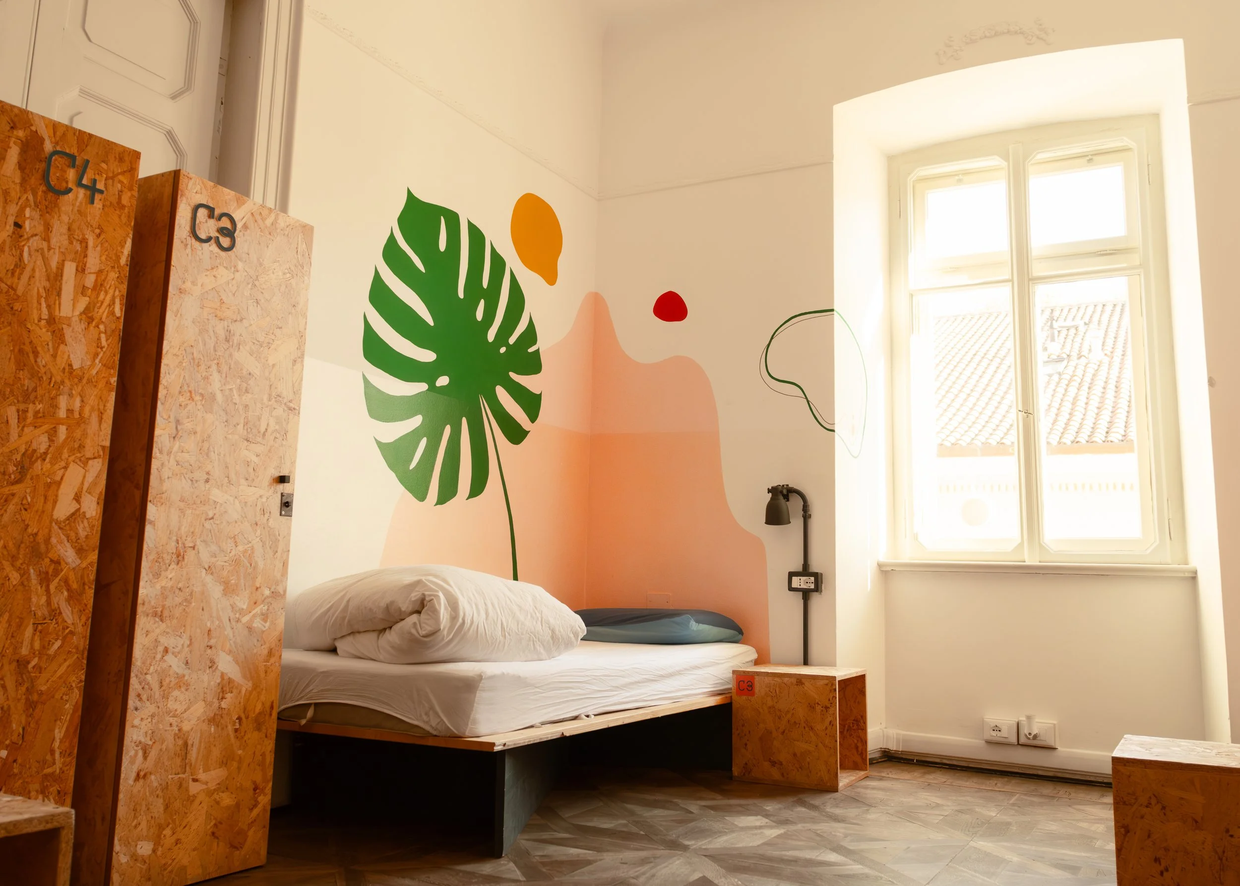 Interior photo of a Controvento Hostel bedroom featuring a green monstera leaf mural, abstract wall shapes, a bed with white linens, and wooden storage boxes labeled ‘C3’ and ‘C4, photographed by Skye J Blount.