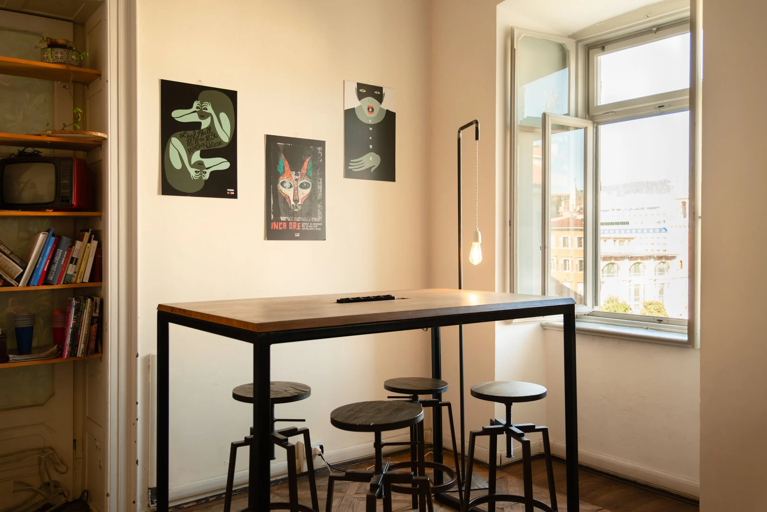 Interior photo of a room corner at Controvento Hostel featuring a wooden high table, four stools, and wall artwork, photographed by Skye J Blount.