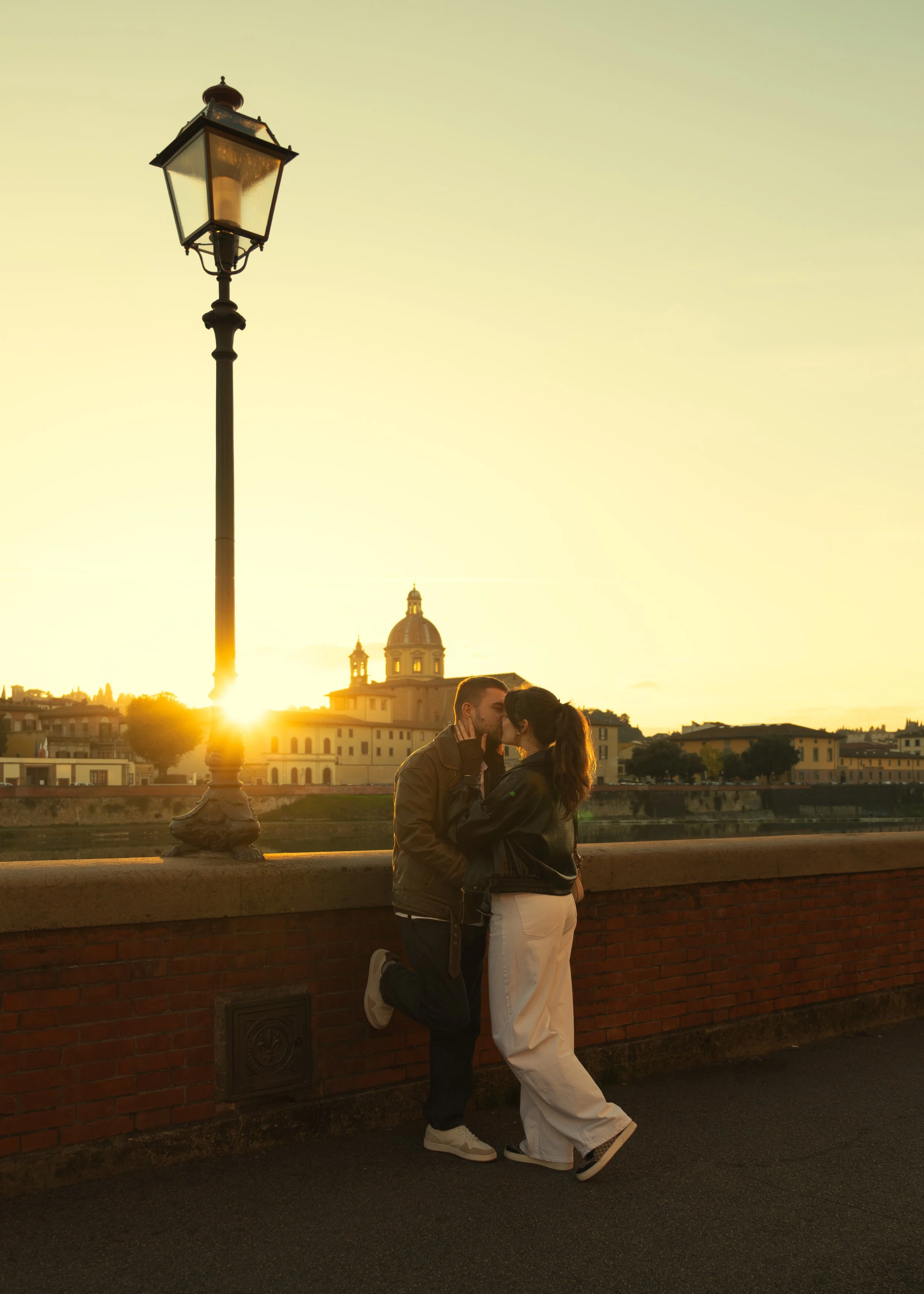 A couple kissing next to a historic cityscape at sunset, with a street lamp and a church in the background.