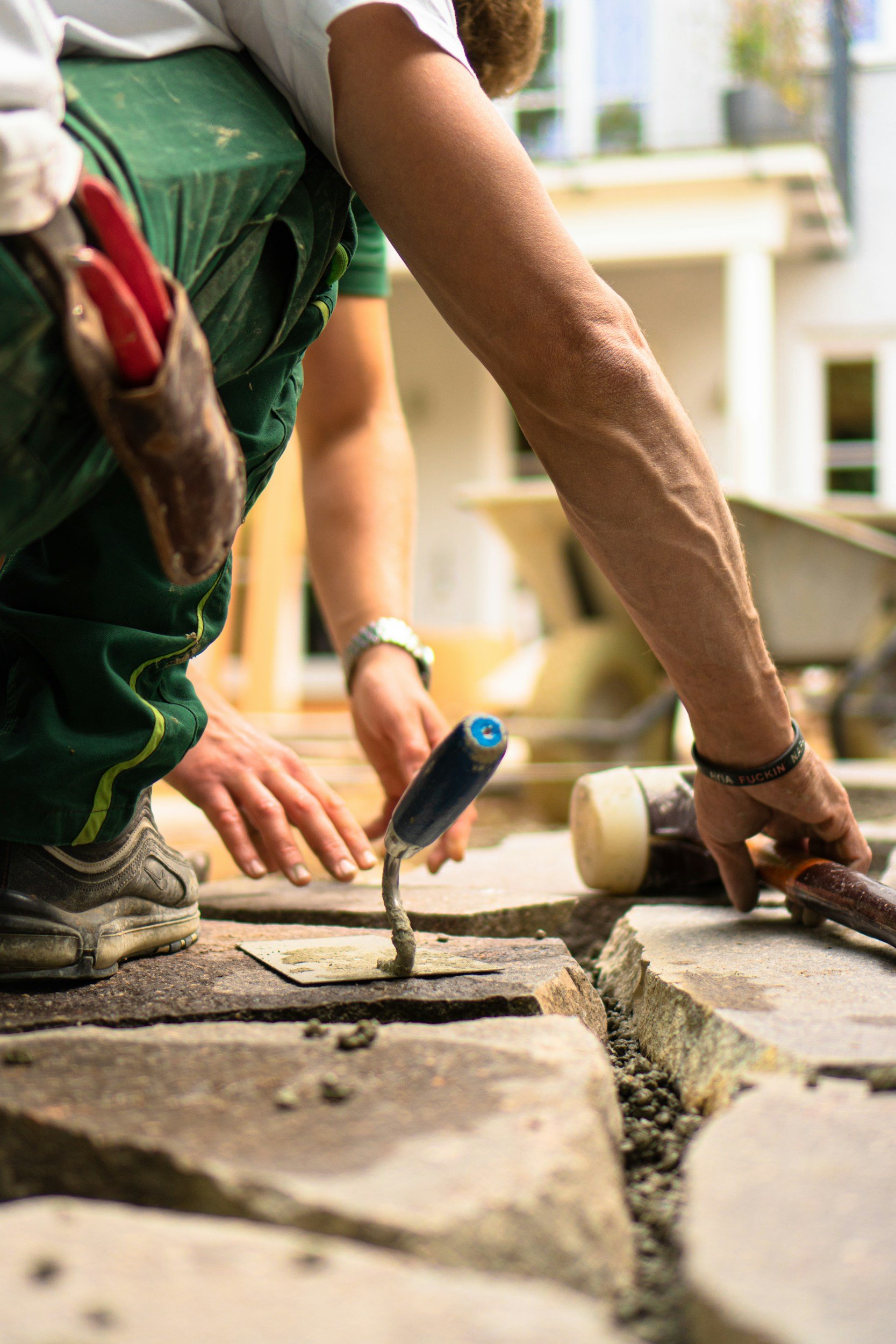 Close-up of a person laying bricks on a pathway, with tools like a mallet and a trowel visible, working on a brick paving project.