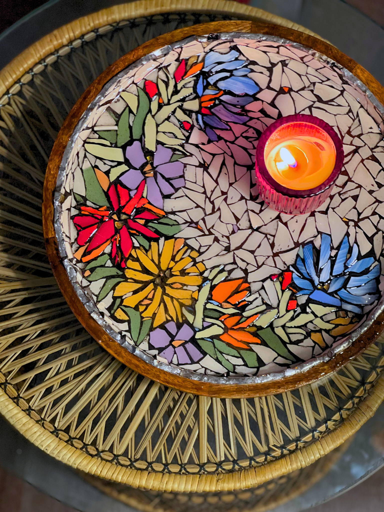 A mosaic tabletop with colorful butterfly and flower designs around a lit candle in the center, placed on a rattan table.