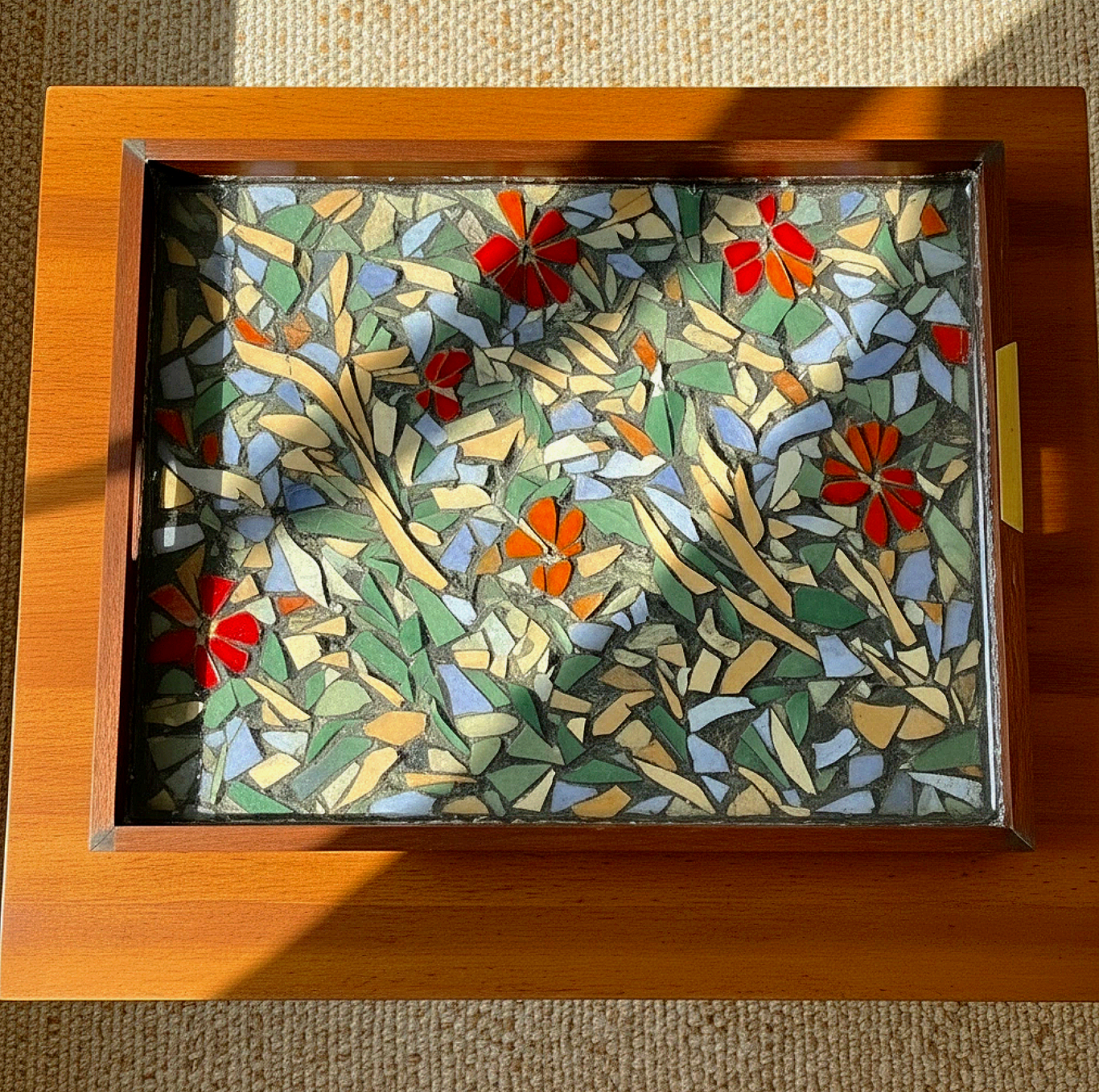 A rectangular mosaic with red, orange, green, and blue floral patterns, set in a wooden frame resting on a wooden surface.