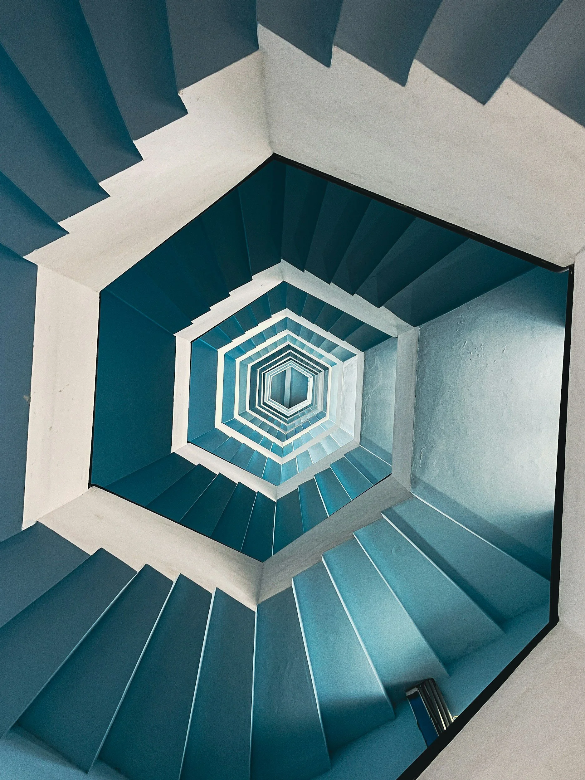 Looking up through a square fluorescent-lit stairwell, the stairs are painted in shades of blue and white, creating a hexagonal tunnel effect.