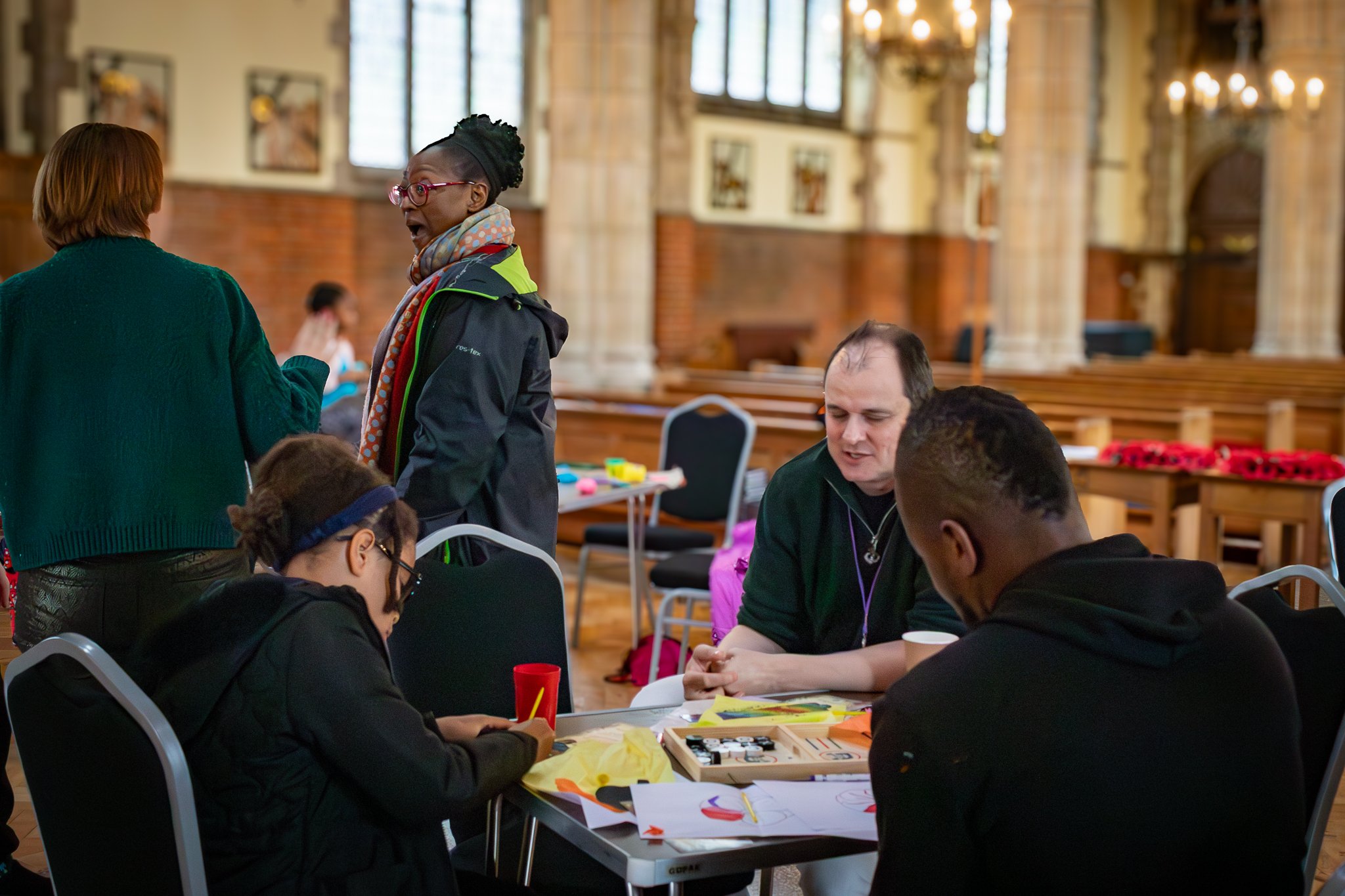 Group of people sitting at a table in a large, ornate room with high ceilings and large windows. Some are engaged in arts and crafts activities, while others are talking.