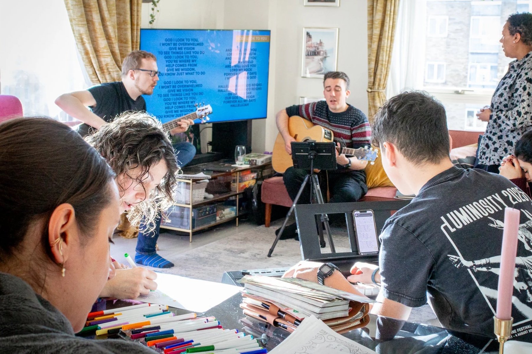 Group of people in a living room, some sitting and some standing, engaging in worship music and singing; two individuals are playing guitars, one singing with eyes closed . A large TV screen displays song lyrics.