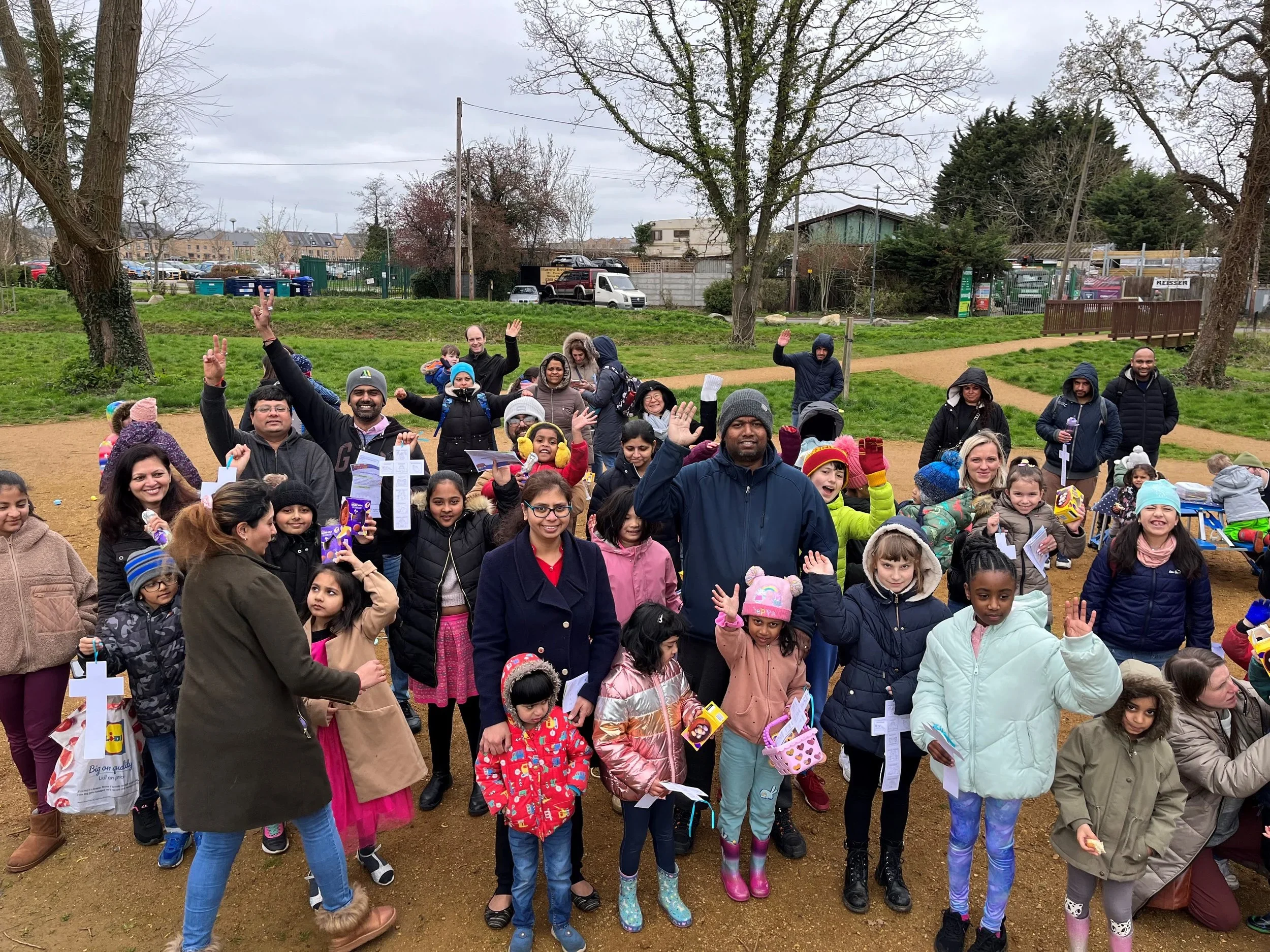 A group of children and adults gathered outdoors in a park on a cloudy day. Some are holding crosses, easter activities and small gifts, and many are waving or smiling at the camera.