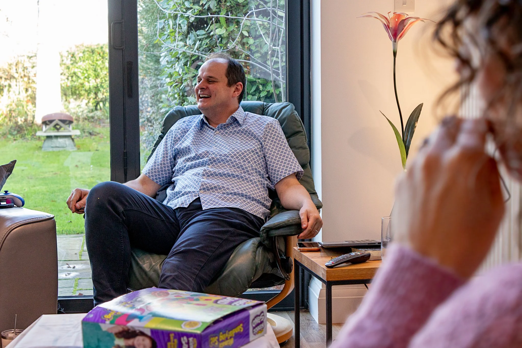Man sitting in a recliner chair, smiling and laughing, near a large sliding glass door with a view of a green backyard.