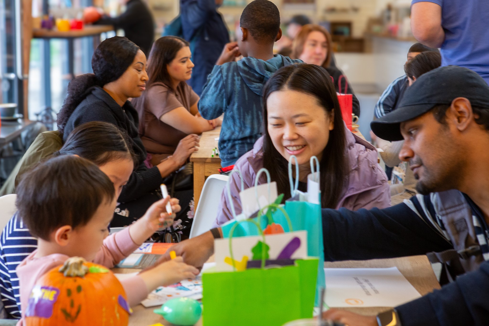 Group of people, including children and adults of various ethnicities, gathered around a table at an indoor event. The table has colorful gift bags and a decorated pumpkin, suggesting a festive celebration.