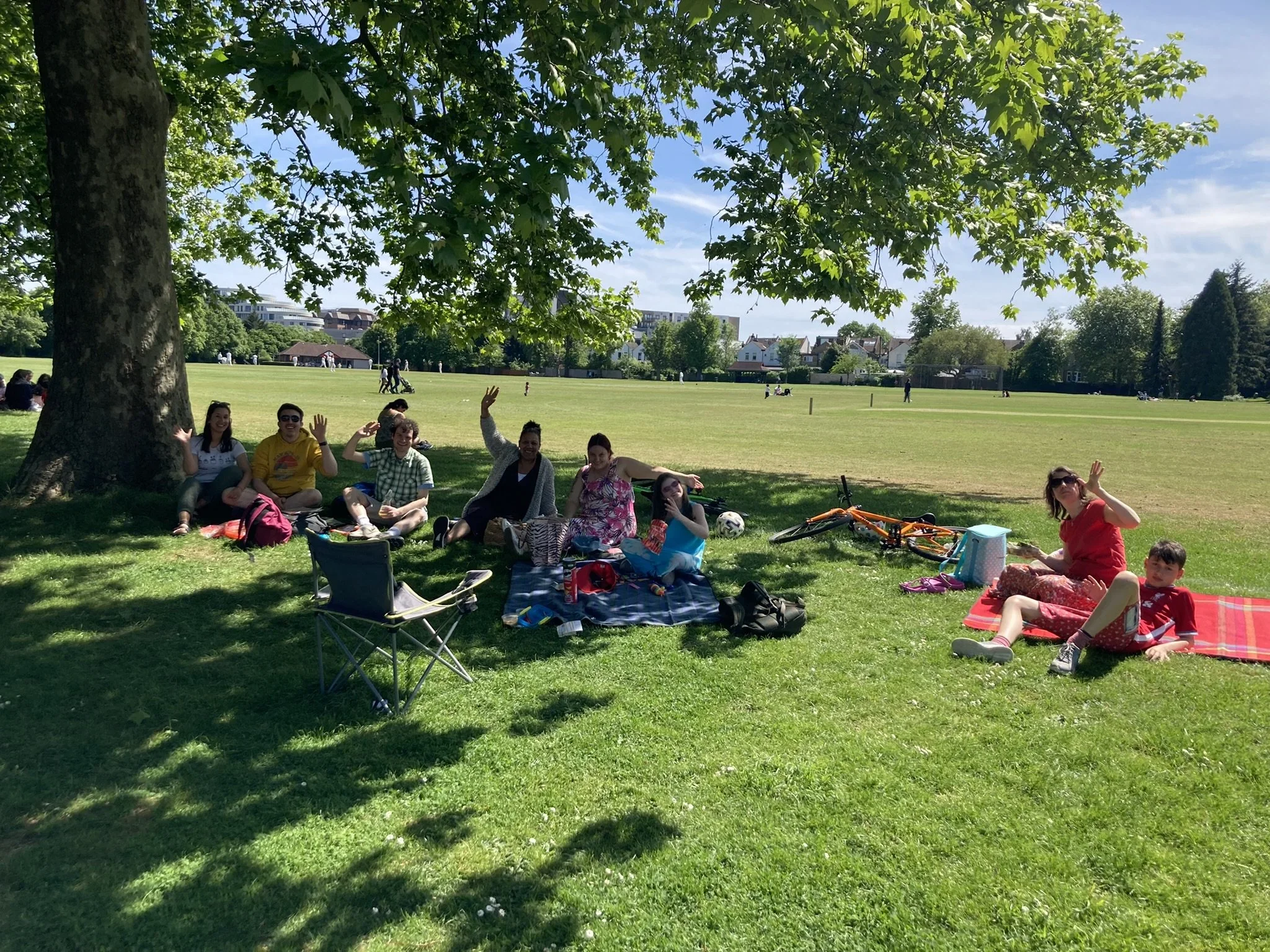 A group of people sitting on blankets under a large tree in a park, smiling and waving at the camera. There are bikes and bags nearby, and a spacious grassy field extends into the distance.