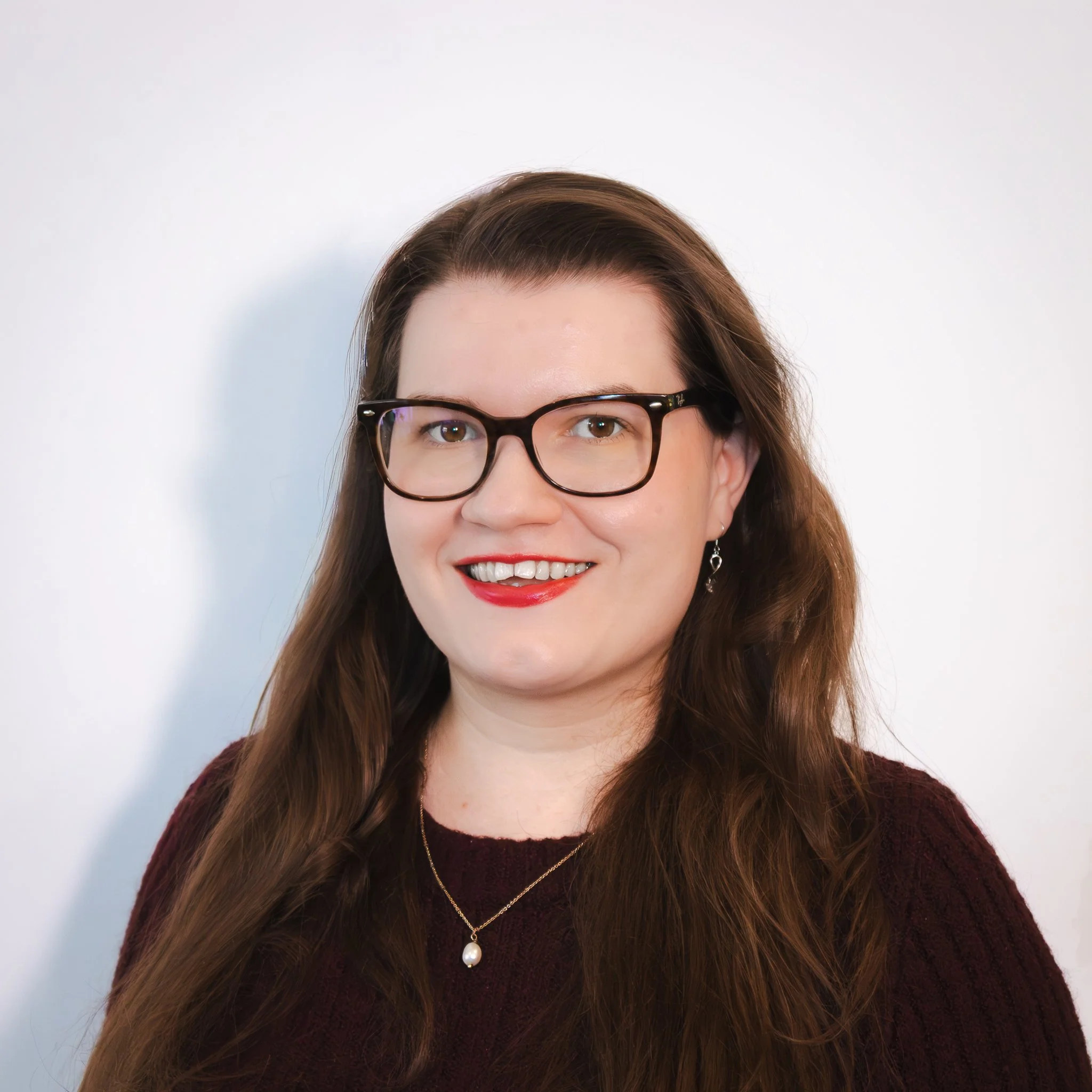 A young woman with long brown hair, smiling, wearing a light-colored top and a pearl necklace, against a plain white background.