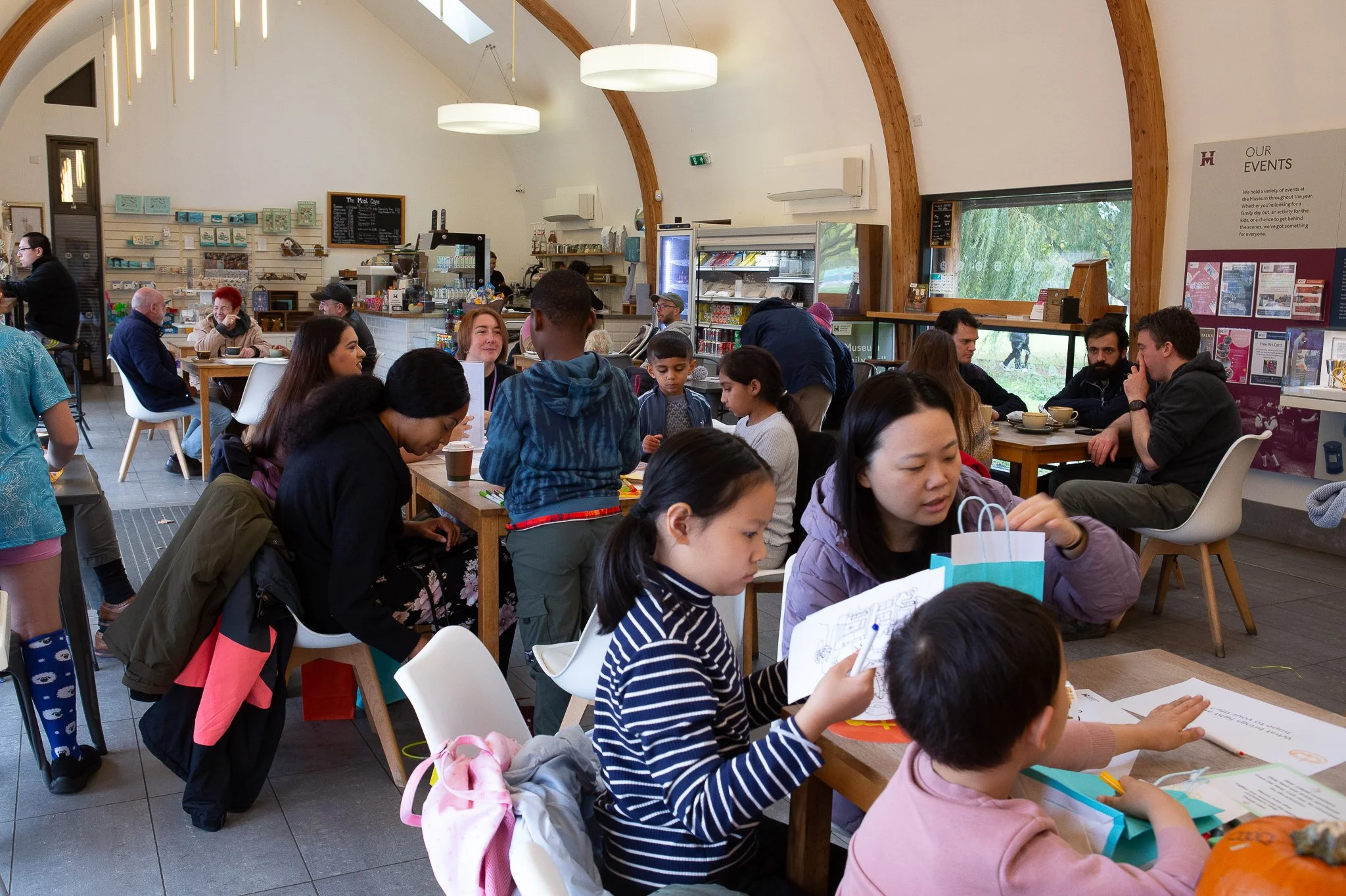 People sitting and standing inside a café with high, wooden arched ceiling, some eating, drinking, and engaging in various activities. The café has a counter, menu board, refrigerator, and posters on the wall.