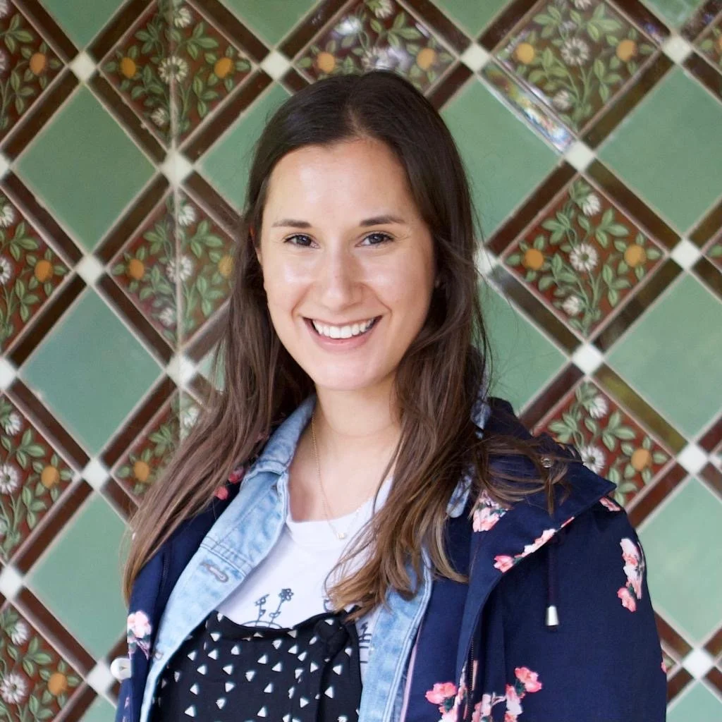 A smiling woman with long brown hair, wearing a denim jacket and a dark blousy shirt, standing in front of a decorative green tiled wall with floral patterns.
