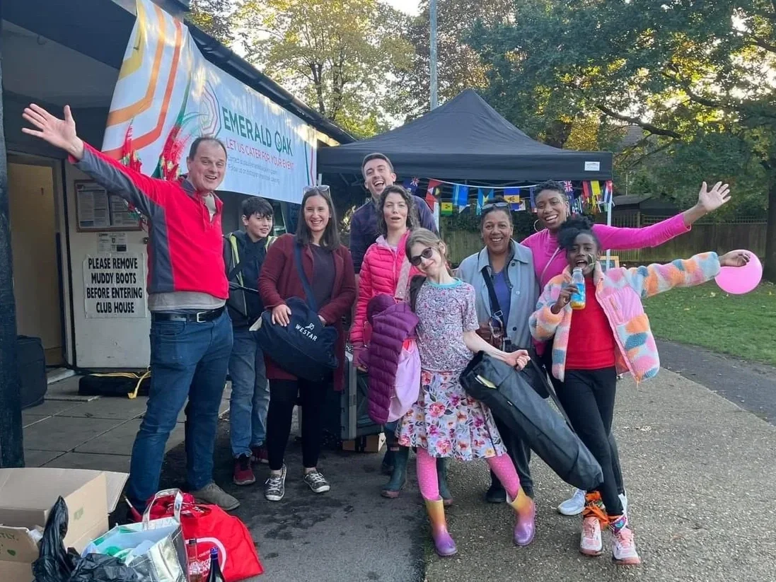 Group of people smiling and waving in front of a black tent at an outdoor community, cultural food event, with colorful bunting, during daytime.