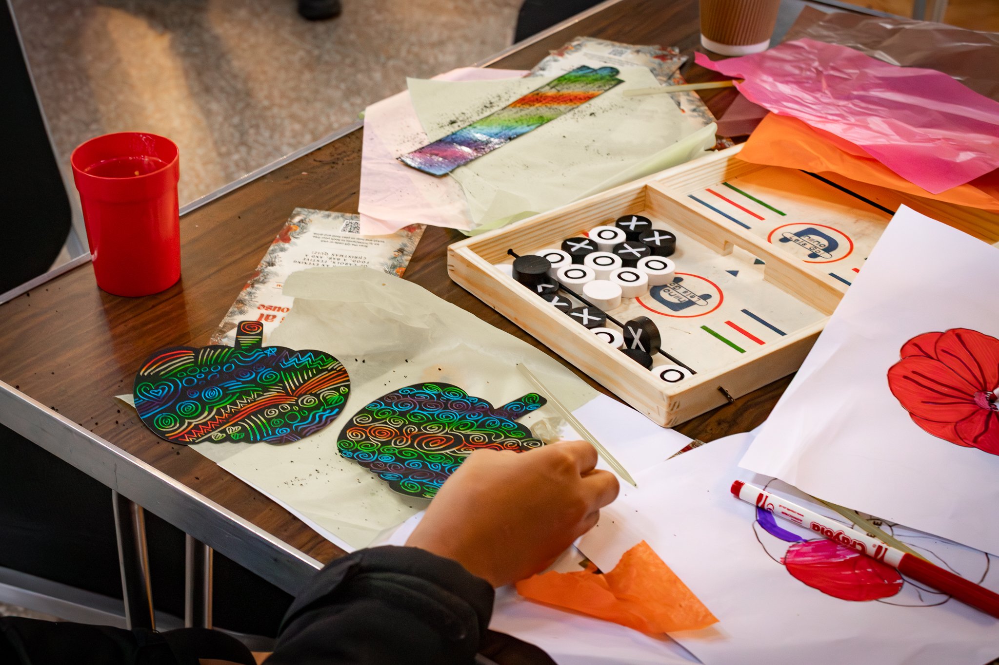 A craft table with colorful papers, a wooden tic-tac-toe game, markers, and rainbow-colored patterned paper cutouts of pumpkins. A person's hand is visible, working on the craft project.