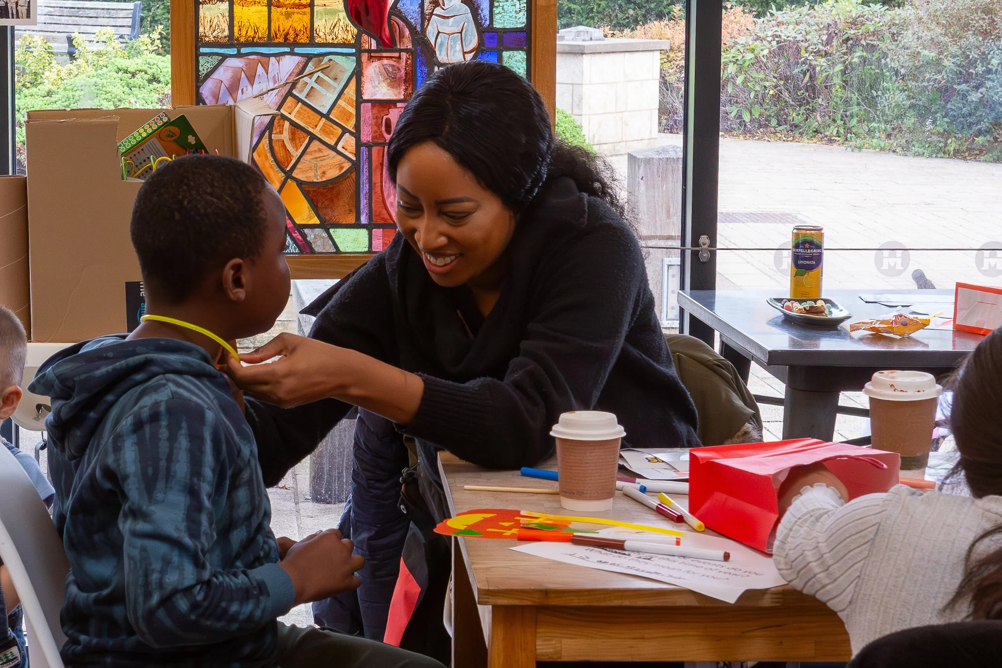 A woman helps a young boy put on a medal around his neck at a table in a room with stained glass art and large windows showing an outdoor patio.