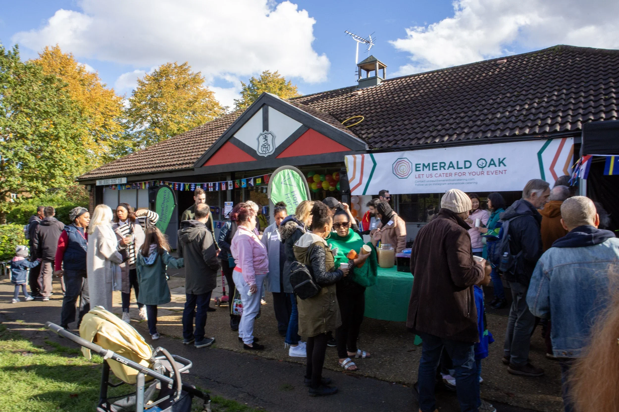 People standing in line at an outdoor event, with a building in the background and a banner with event details. The scene is set on a sunny day with trees showing fall foliage.