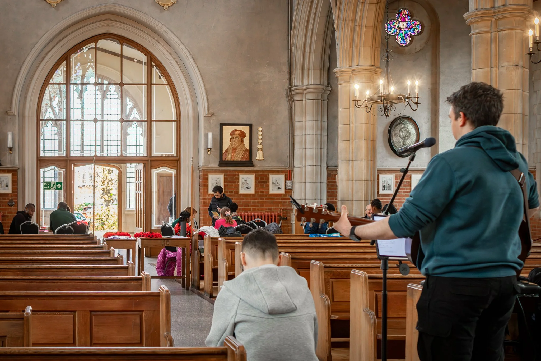 People inside a church-like building, some sitting and some standing, while a man plays worship in the guitar and sings into a microphone, with stained glass windows and religious artwork on the walls.