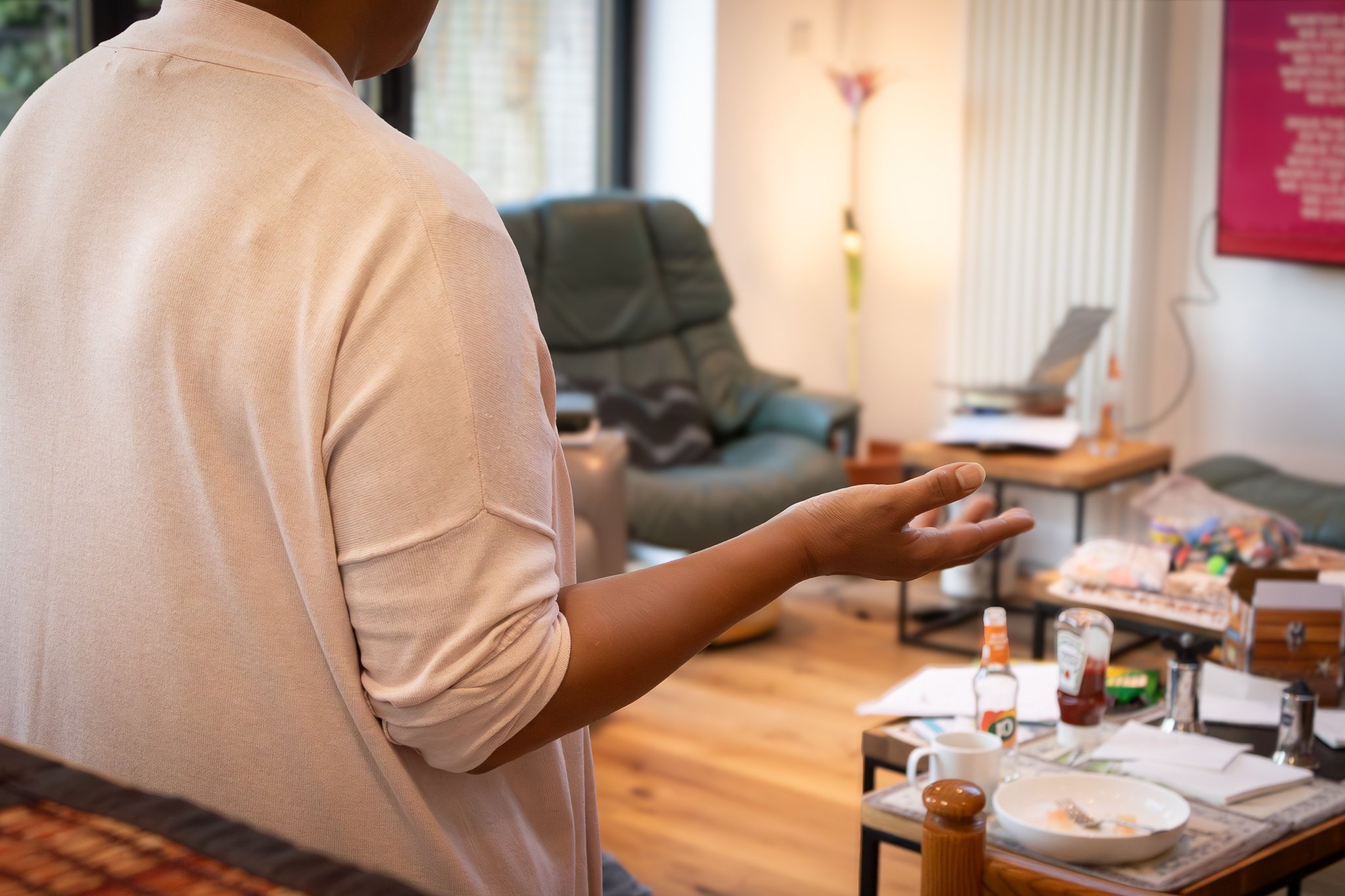 Person in a beige top gesturing in a cluttered living room with a table with bottles, cups, and papers, and a couch in the background.