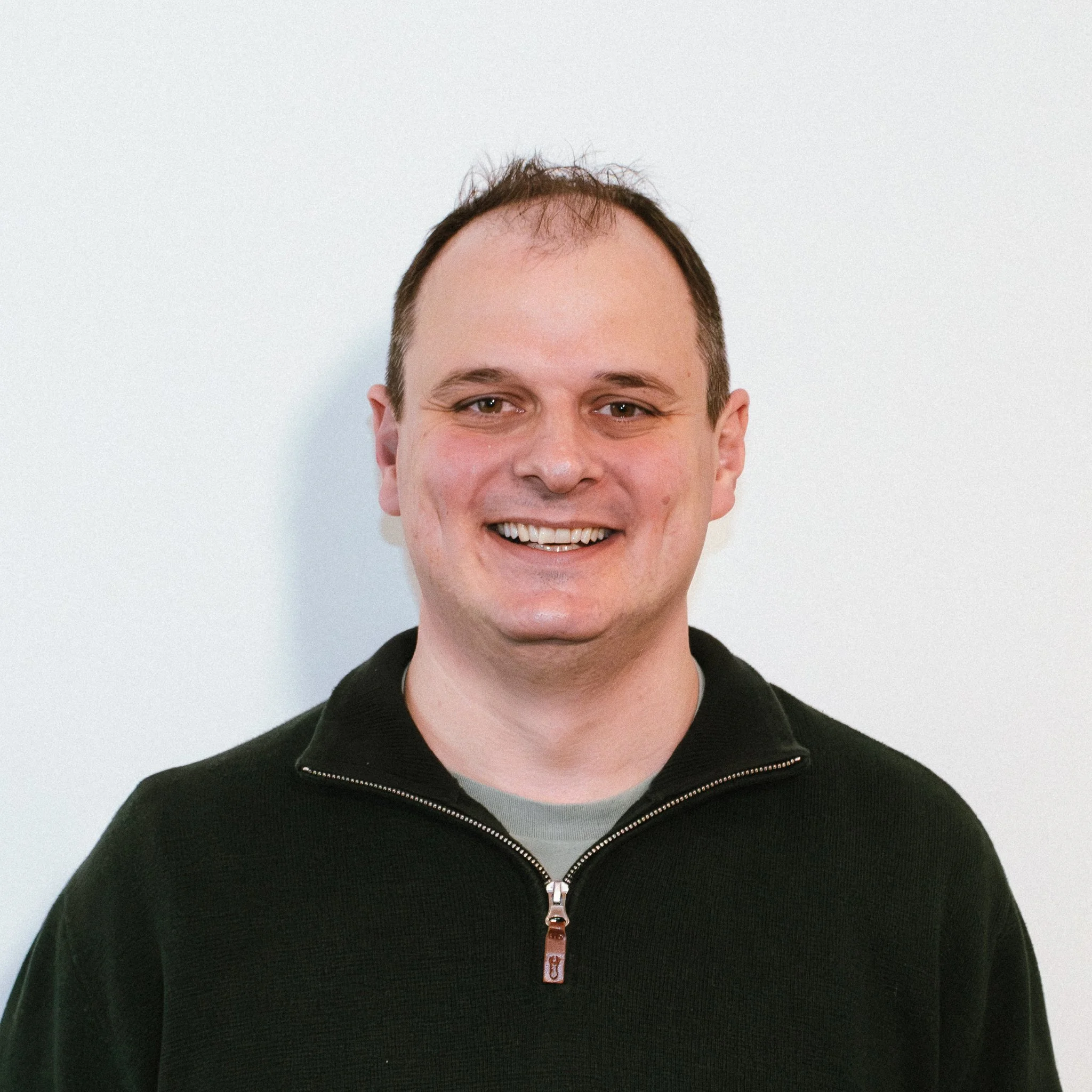 A man with a cheerful smile leaning on a counter, wearing a festive red sweater with a reindeer face, inside a small shop or kiosk.