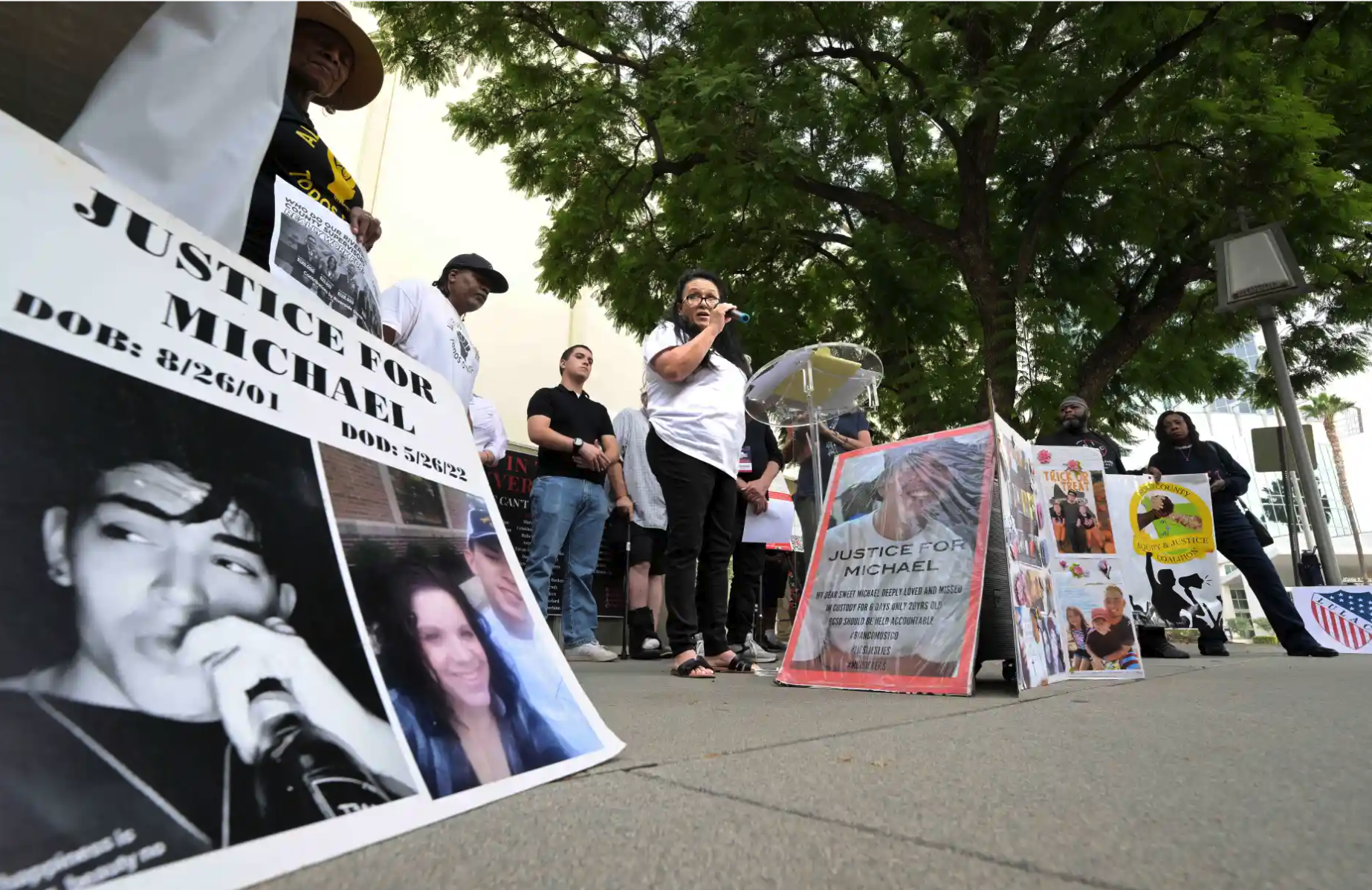People gathered at a vigil or protest holding signs and pictures advocating justice for Michael Vargas, with Kathy Nigro (mother) speaking at a microphone. Signs include a photo of Michael and personal messages demanding accountability for his death.