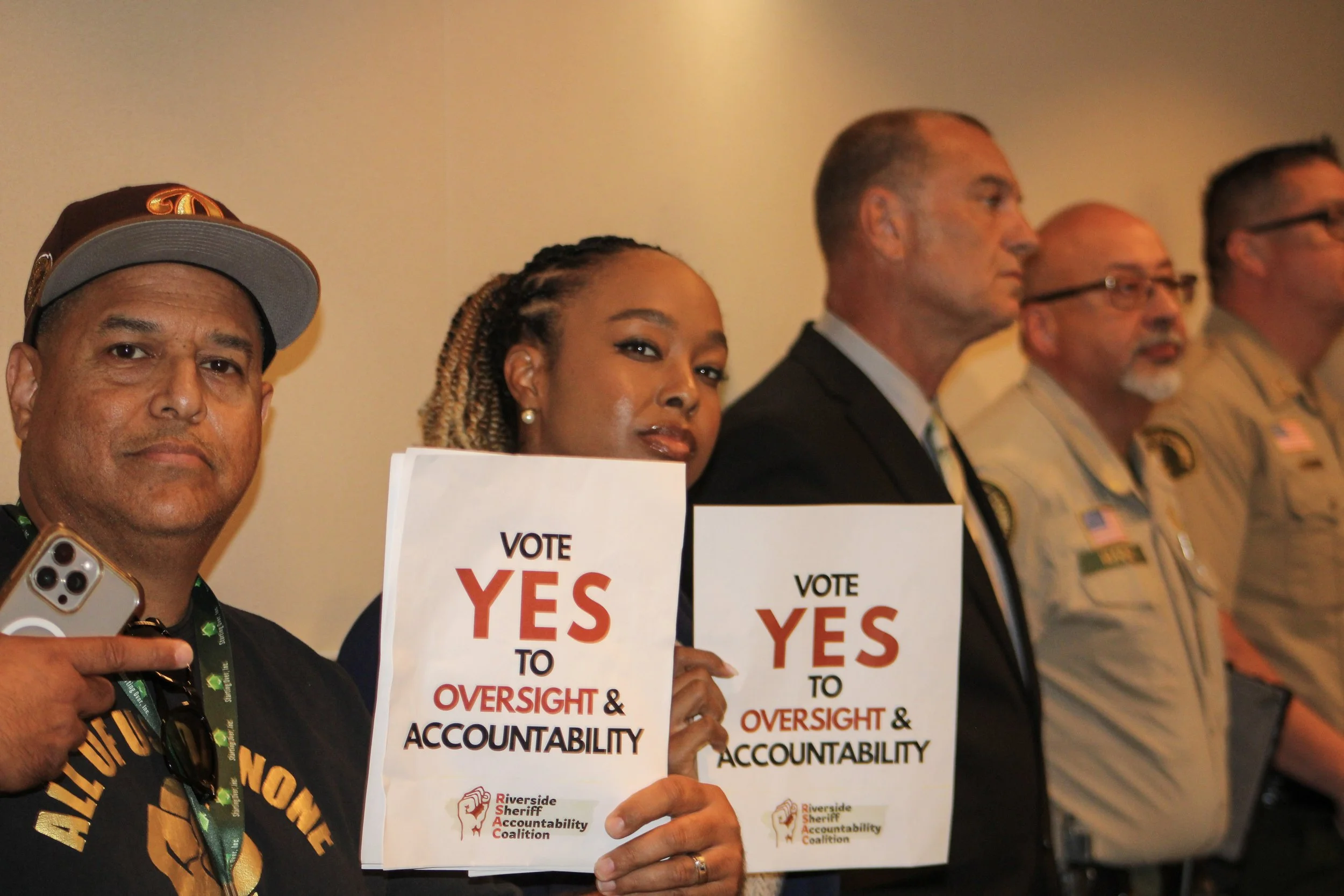 Group of diverse individuals holding signs that read 'Vote Yes to Oversight & Accountability' at a rally or meeting.
