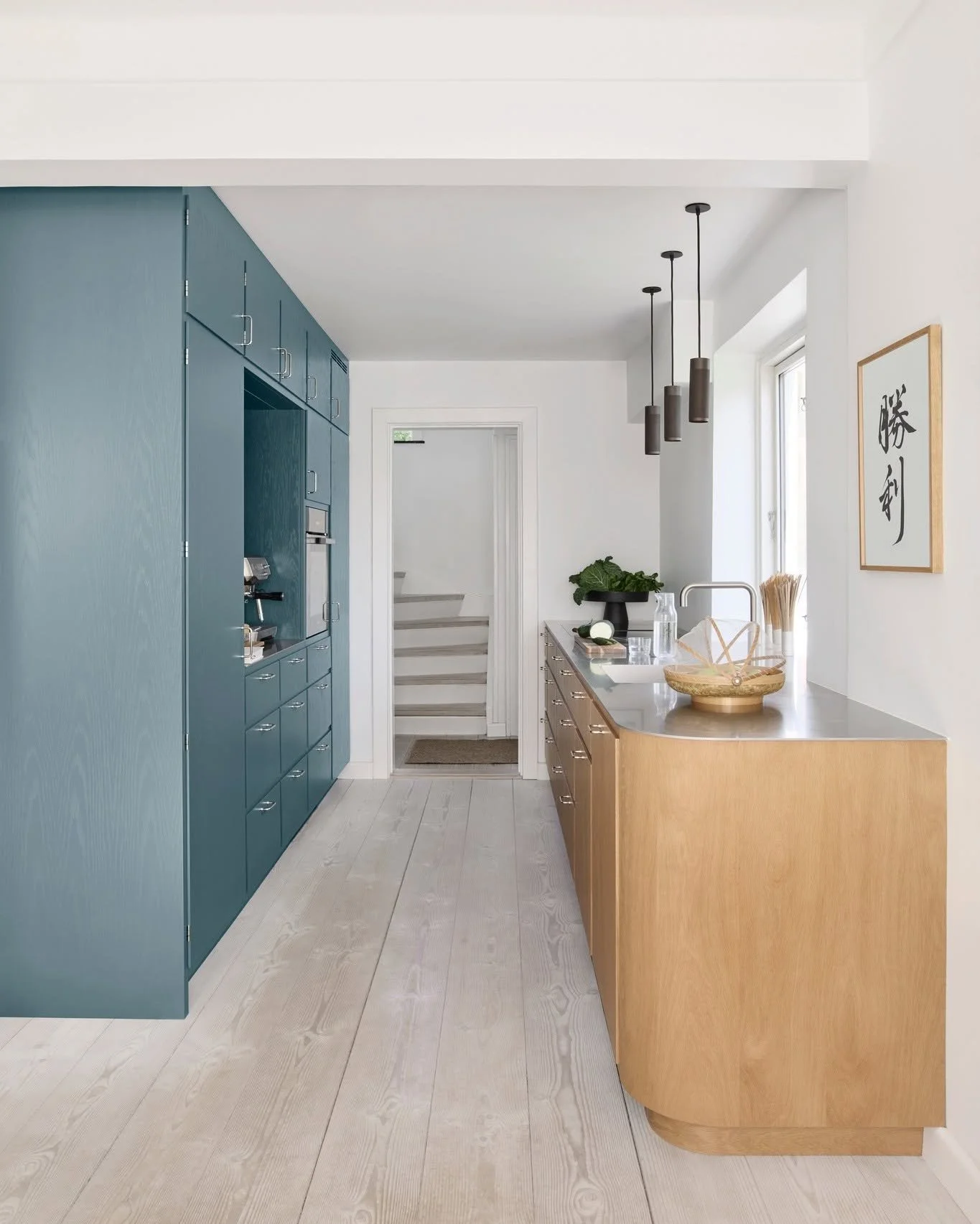 Interior view of a modern kitchen with blue cabinets on the left, a wooden kitchen island on the right with a sink, pendant lights above, a window, a framed artwork with Japanese characters on the wall, and stairs in the background.