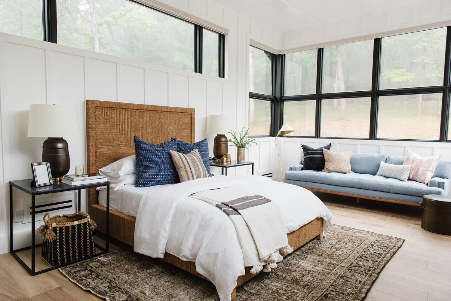 A bright bedroom with large windows showing a wooded outdoor view, featuring a bed with a rattan headboard, white bedding, navy and striped throw pillows, two black nightstands with lamps and decorative items, a light blue sofa with multiple pillows, a floor lamp, and a patterned area rug.