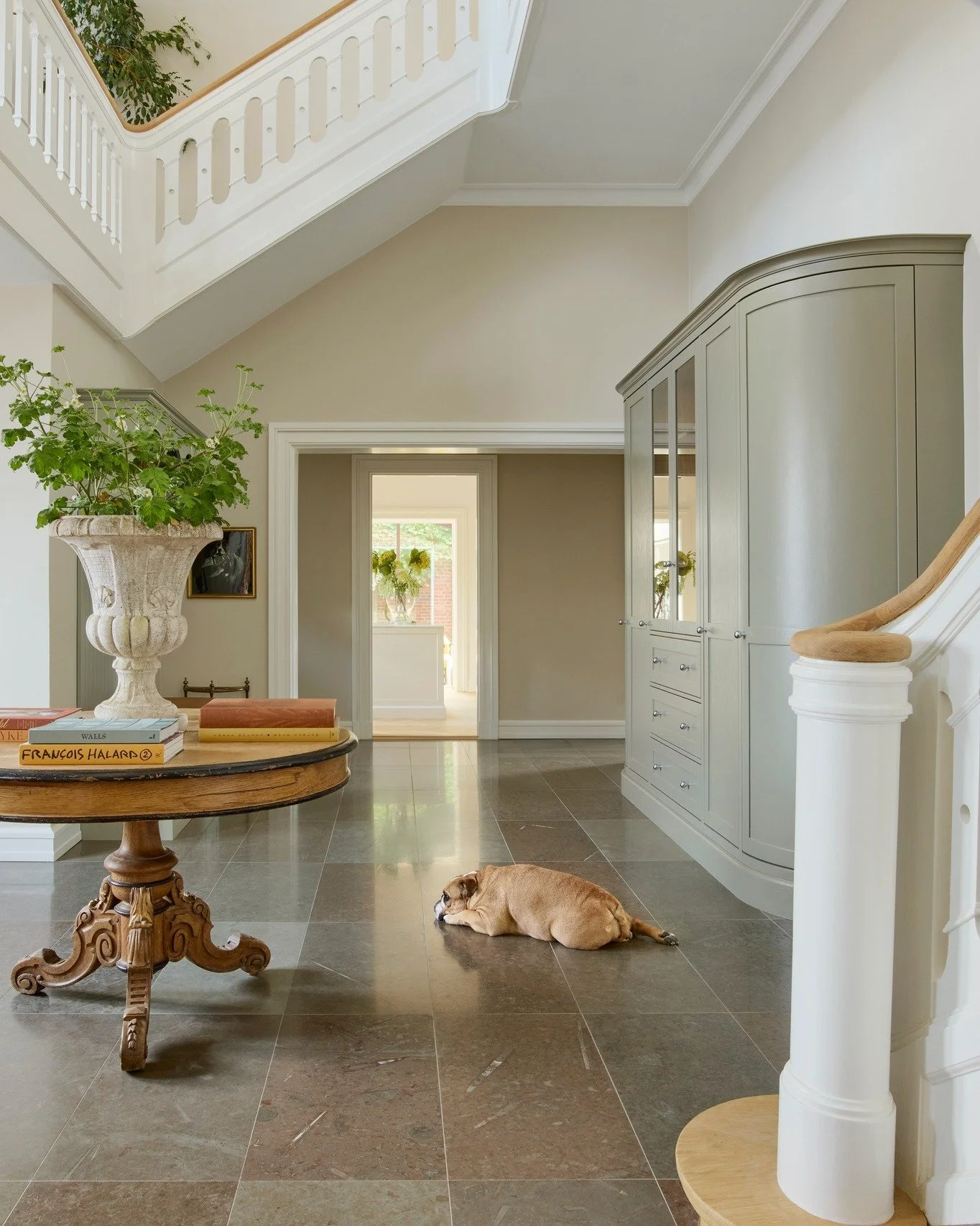 Interior of a house hallway with beige walls, gray cabinet, stone tile floor, a dog lying down on the floor, and a round wooden table with books and a large vase with green plants.