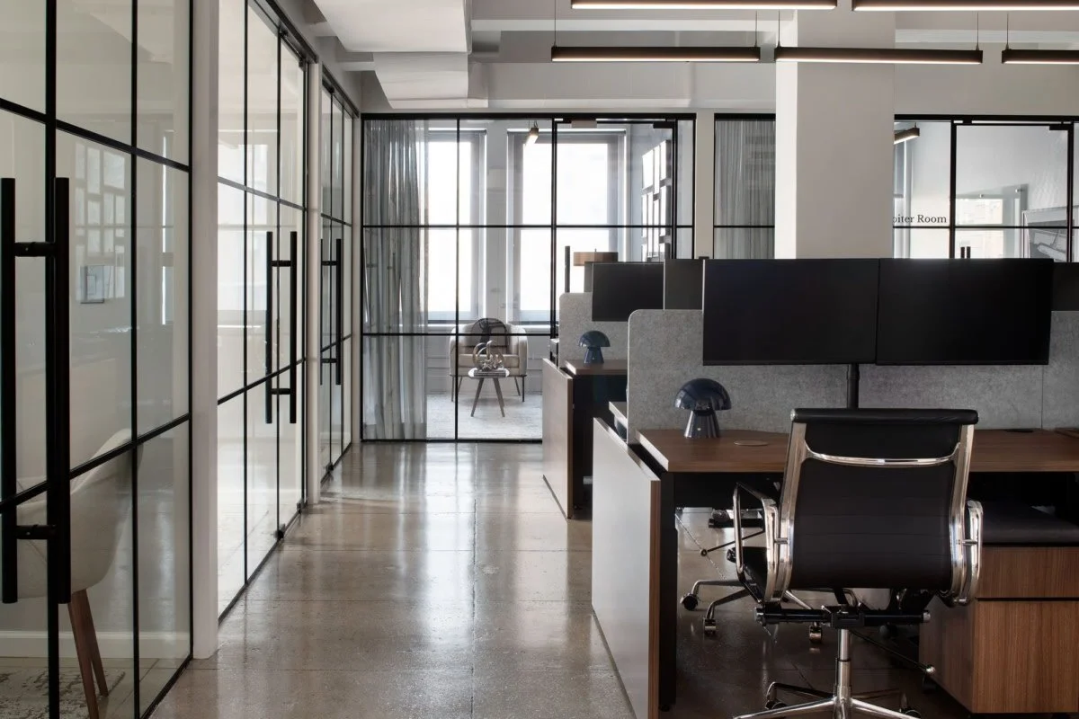 Modern office space with glass-walled conference rooms on the left, open work desks with black desktops, gray fabric partitions, and black office chairs with chrome accents. In the background, a small seating area with a chair, a small table, and a large window with curtains. The floor is polished concrete.