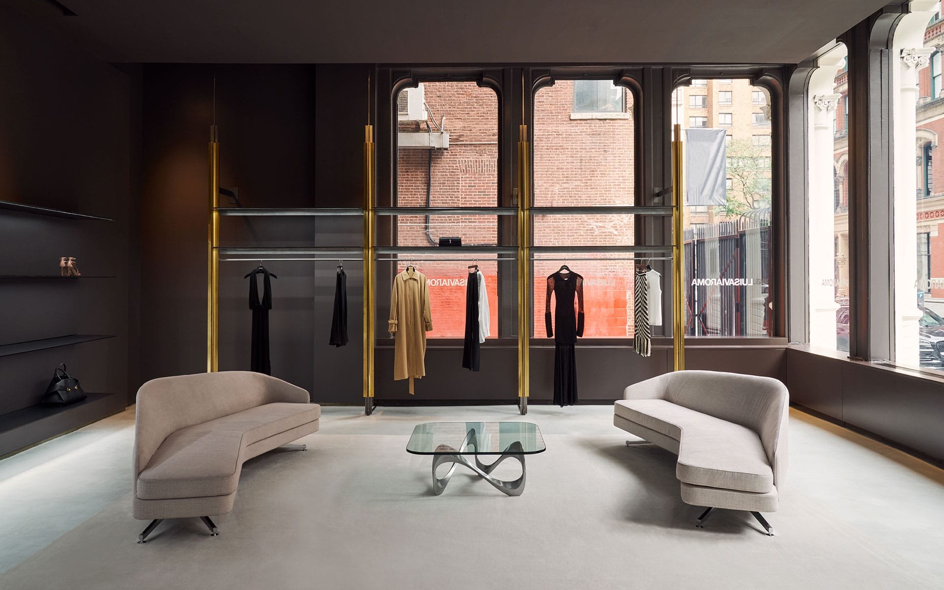 A modern boutique with large windows, two beige curved sofas facing each other, a glass coffee table in the center, and clothing racks with garments hanging in the background.