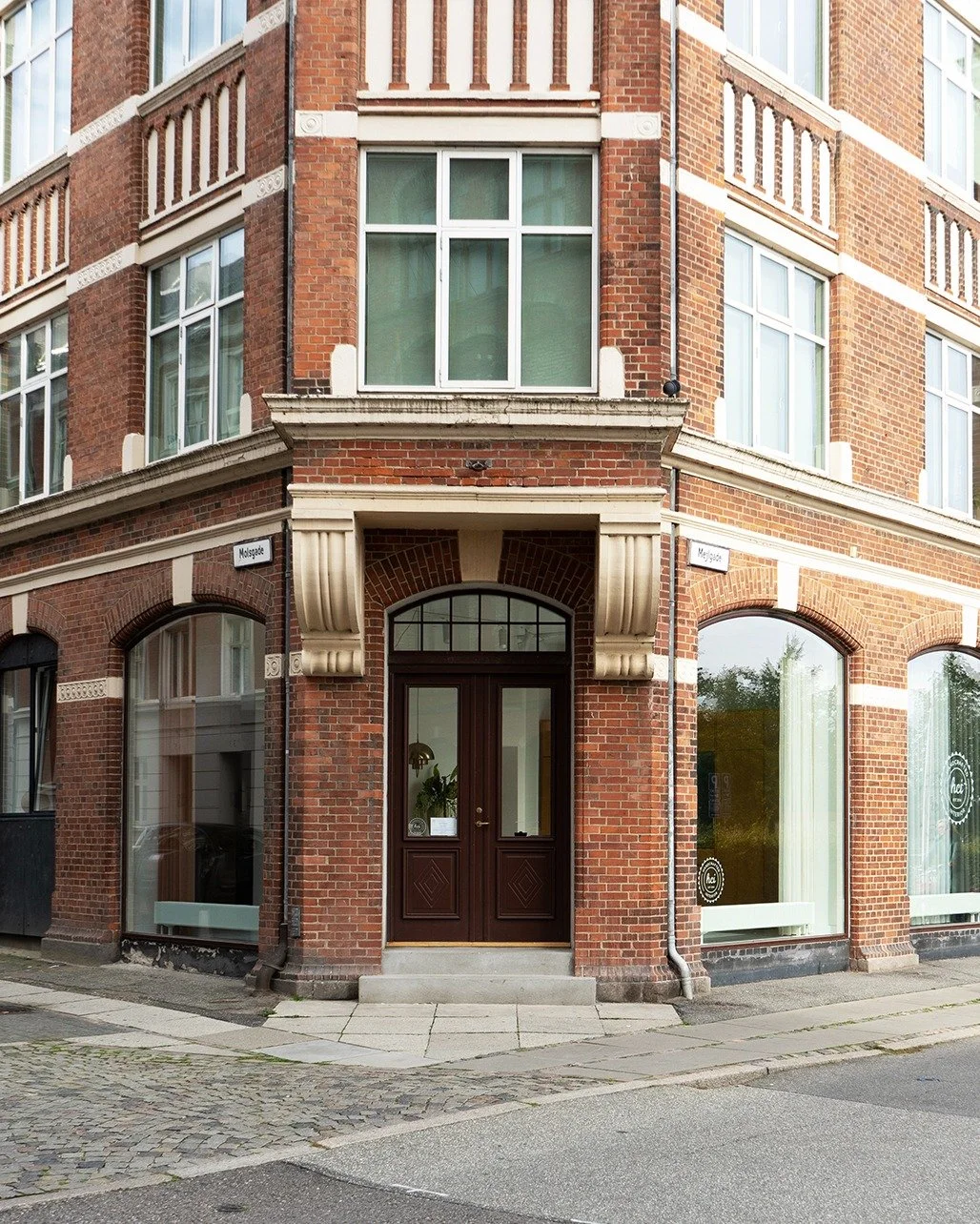 Corner of a brick building with large windows, a central wooden door, and decorative architectural elements at the entrance, located at the intersection of two streets.