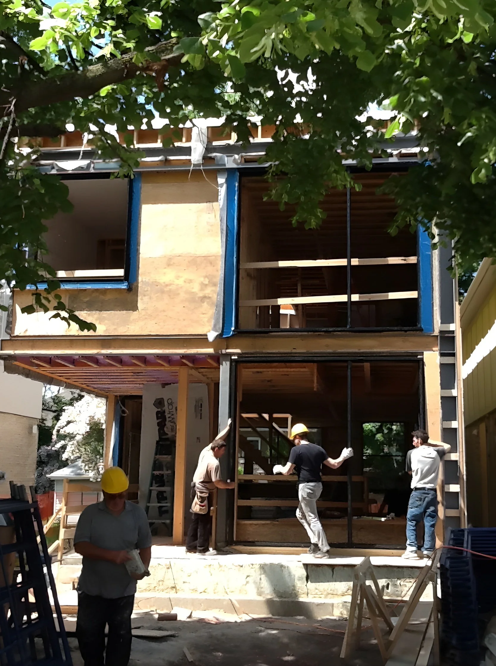 Construction workers working on a multi-story house under construction, with some workers wearing yellow helmets and construction gloves, and the house's framing and exterior walls being built.