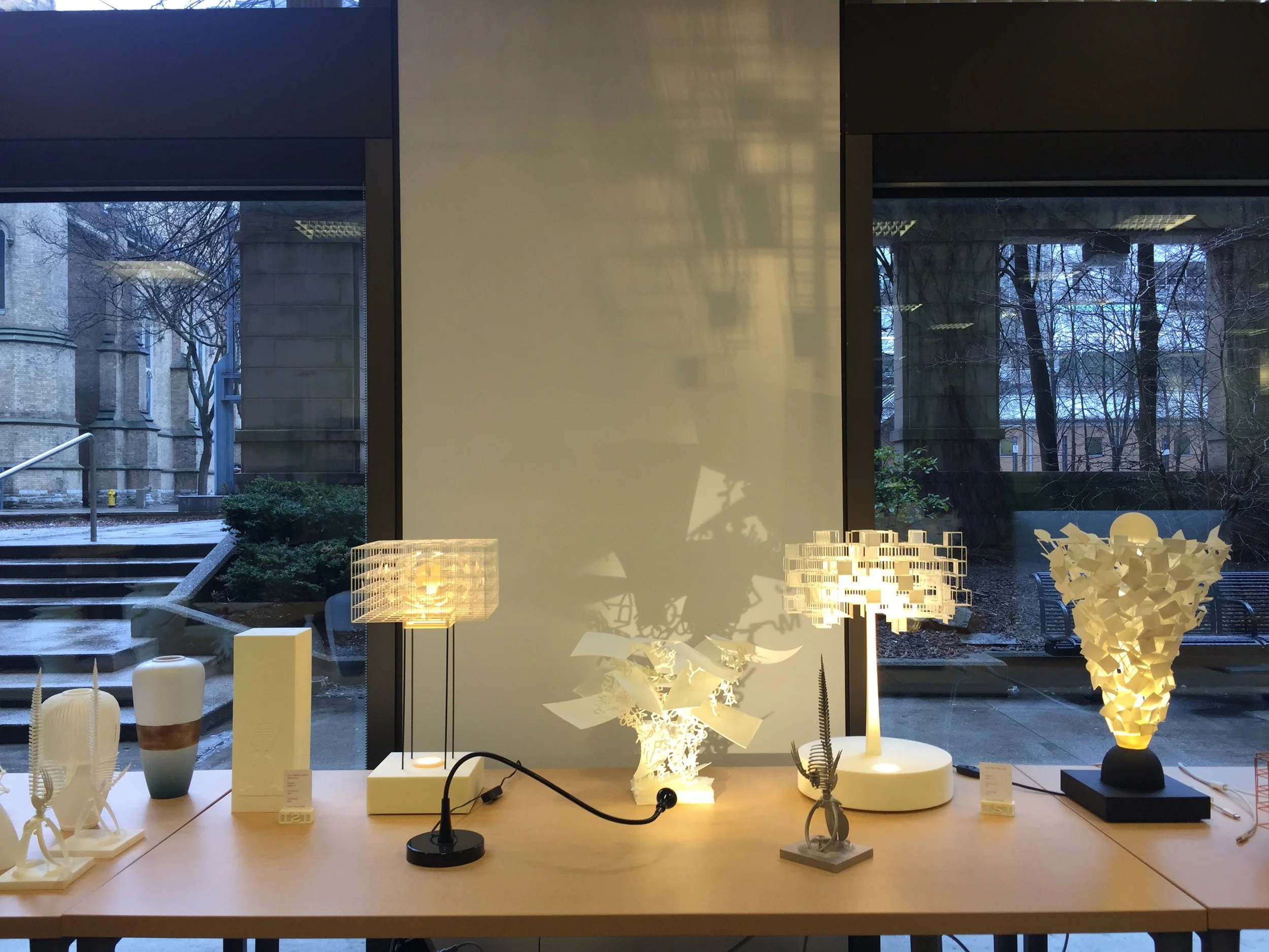 Display of 3D generated decorative white table lamps in a showroom with large windows showing outdoor trees and snow outside.